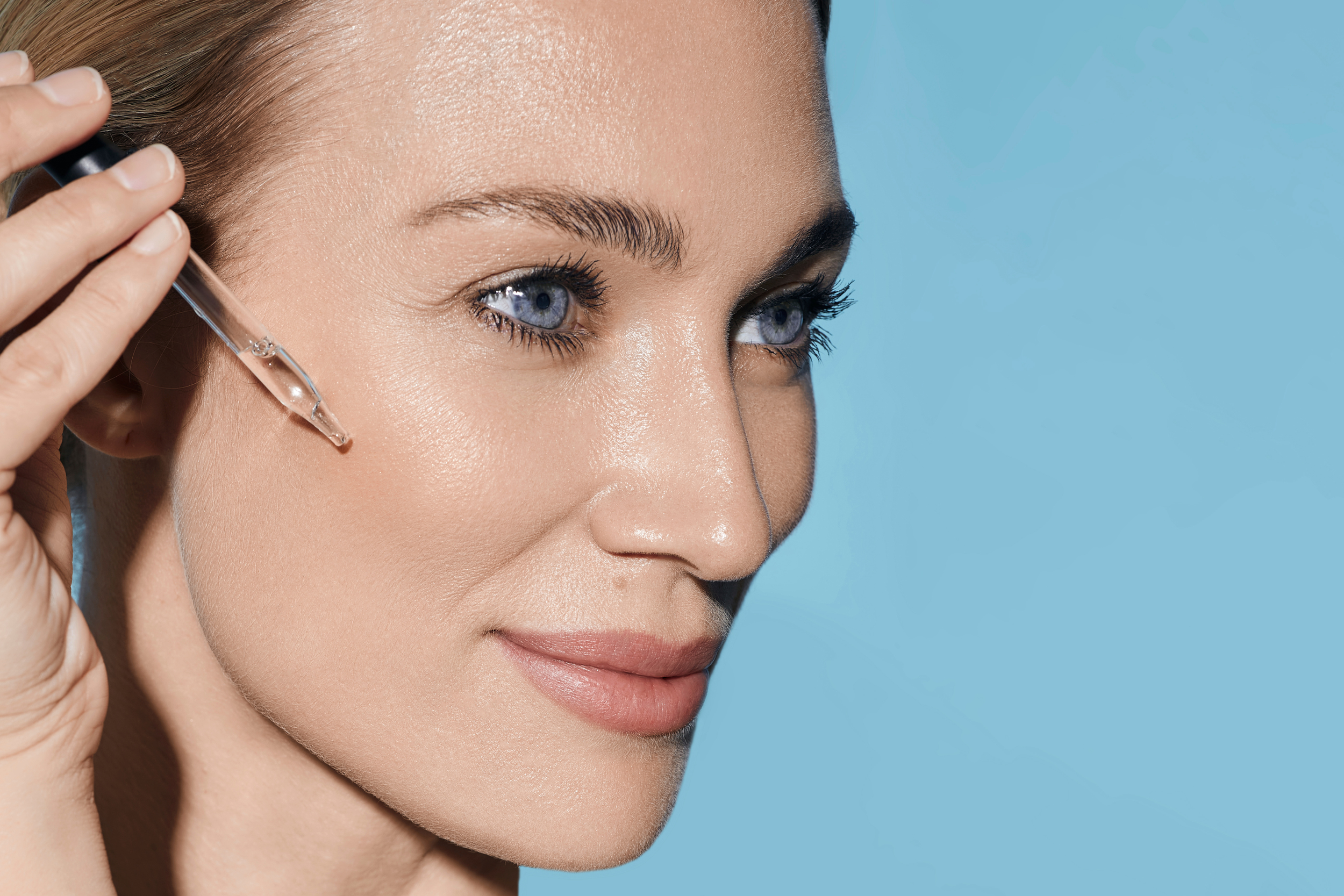 Woman applying skincare serum with dropper on face against blue background.