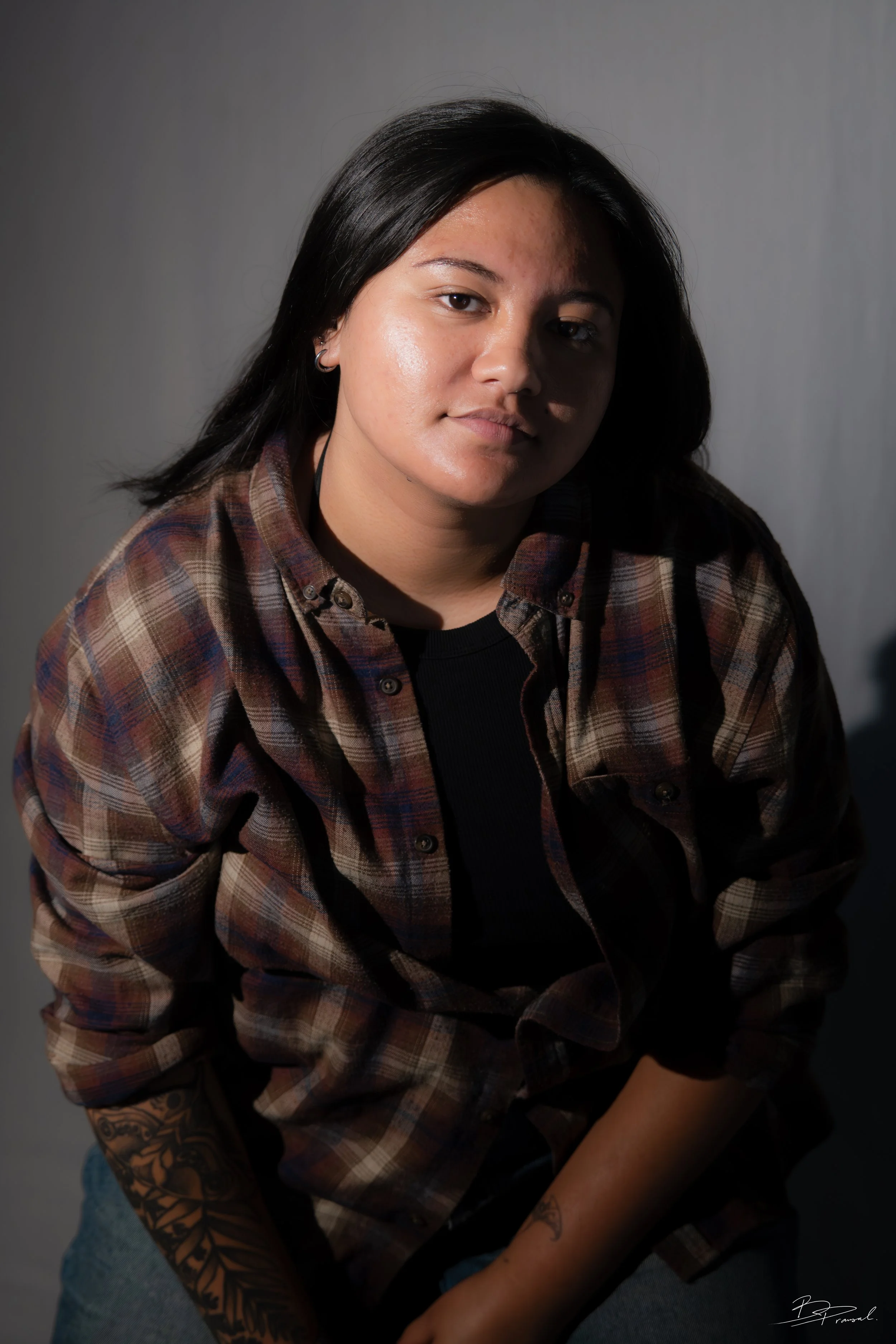A young woman with black hair and tattoos on her arms, wearing a brown plaid shirt over a black top, sitting against a plain background.