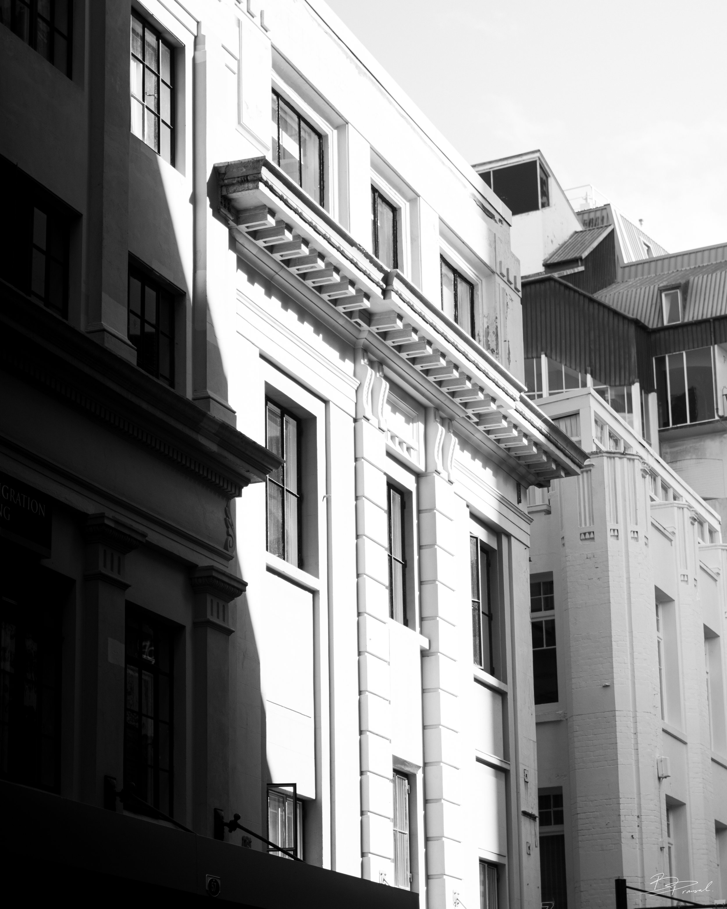 Black and white photograph of multi-story urban buildings with ornate architectural details, window styles, and shadows cast by sunlight.