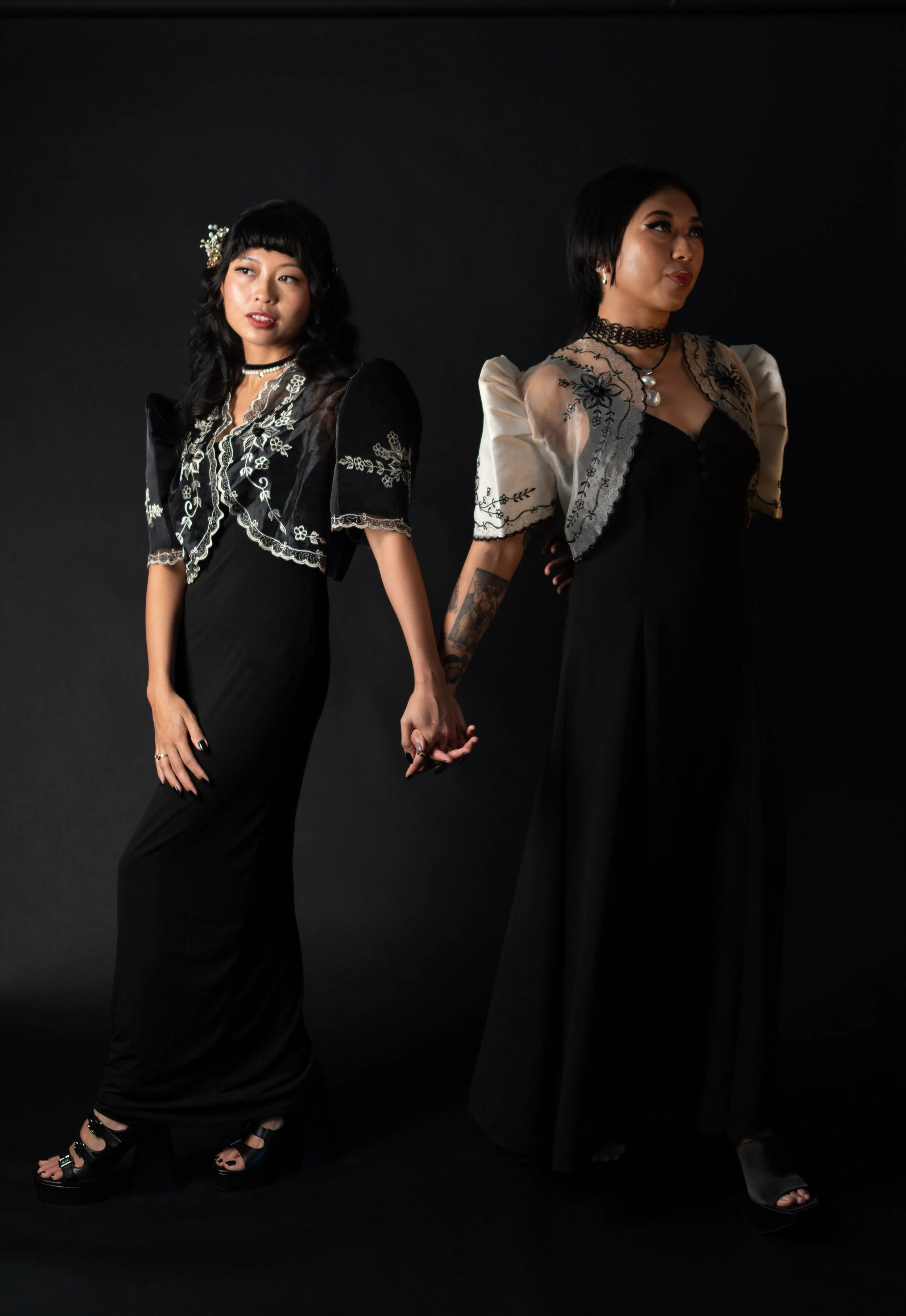Two women dressed in black and cream dresses holding hands against a black background.