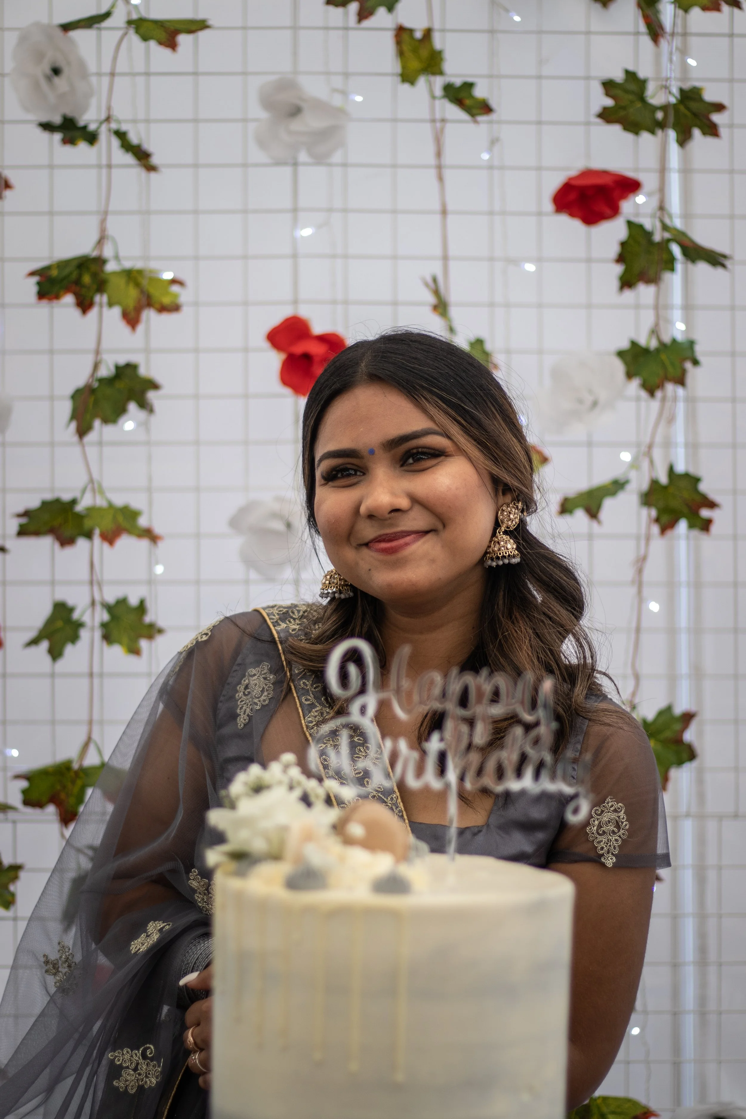 Smiling woman in traditional Indian attire standing behind a white birthday cake with a 'Happy Birthday' topper, decorated with flowers and treats, against a backdrop with green leaves, white and red flowers, and small lights.