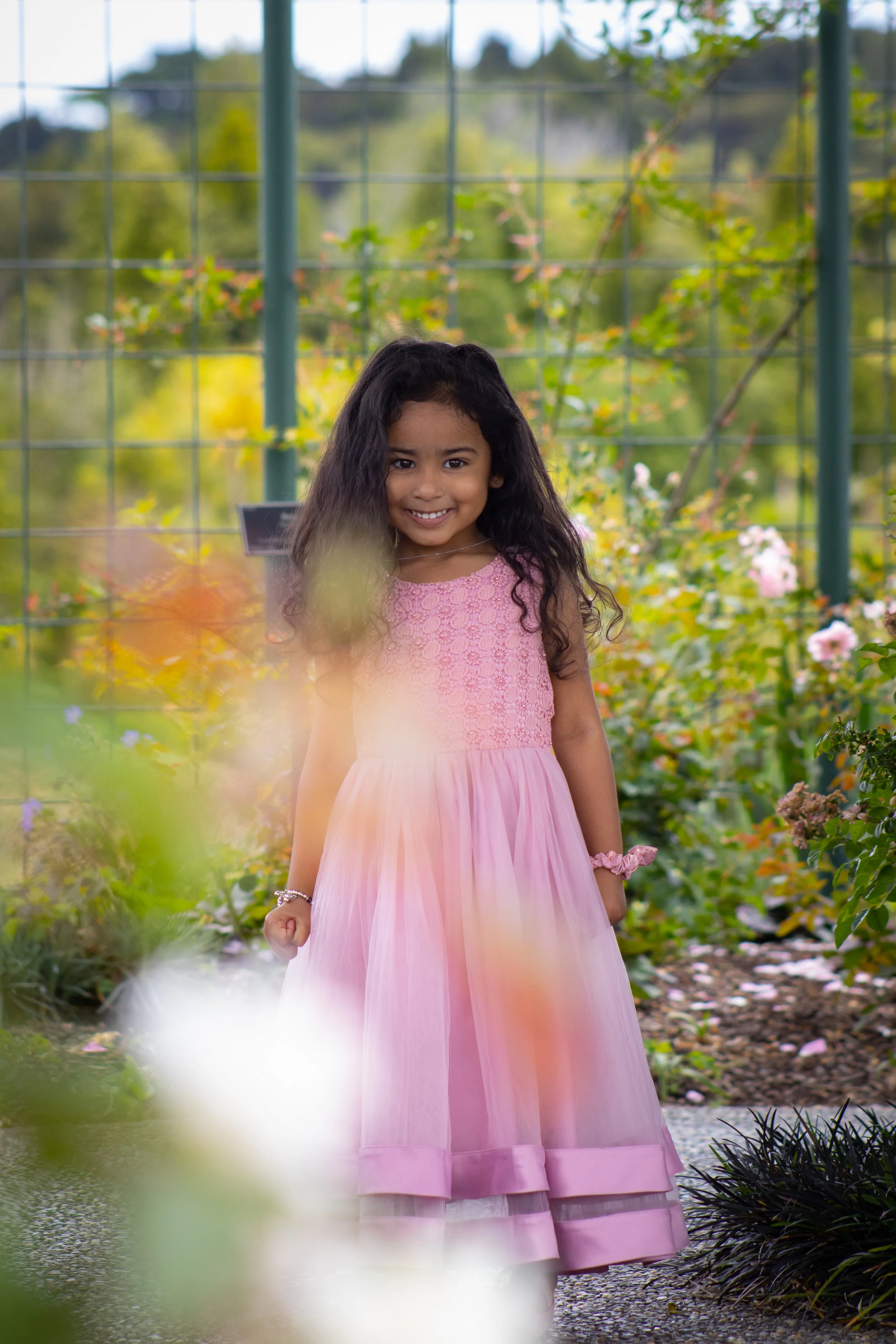 A young girl with long dark hair and a pink dress smiling in a garden with flowers and greenery.