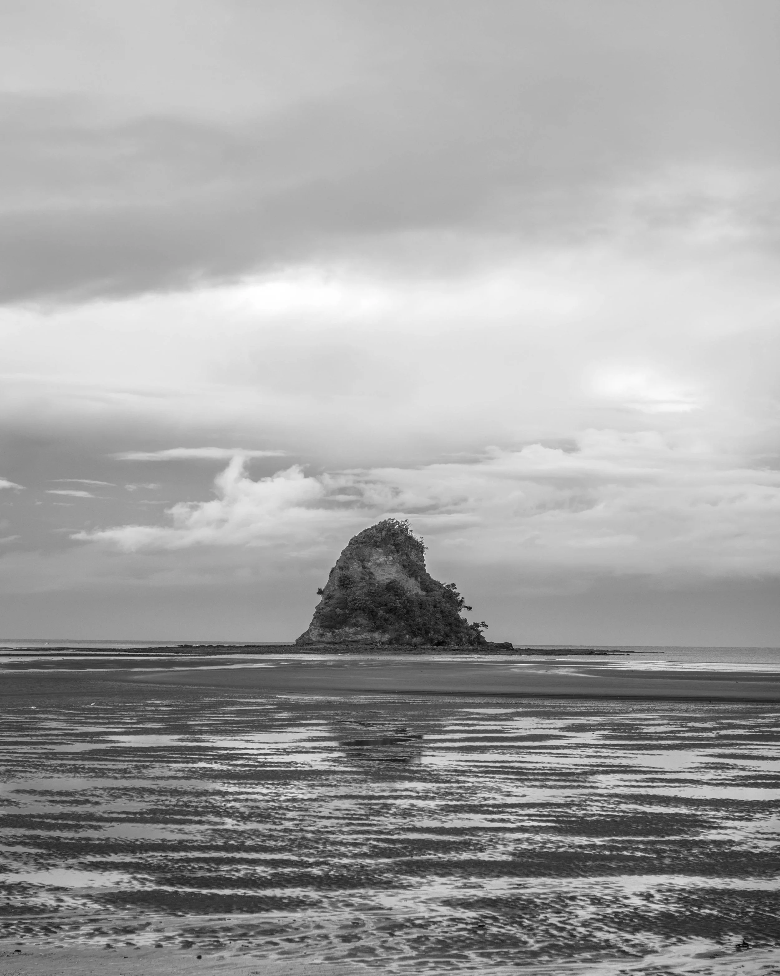 A black and white photo of an island with a rocky, tree-covered summit in the ocean, with an overcast sky and rippled water in the foreground.