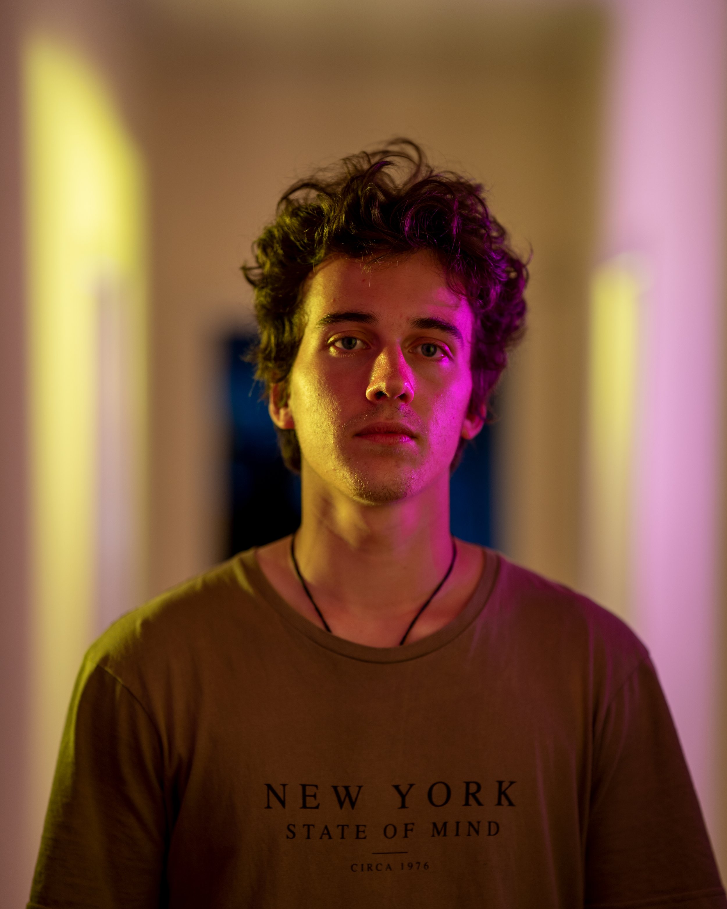 Young man with curly hair wearing a beige T-shirt with text, illuminated with purple and yellow lighting, standing in a hallway.