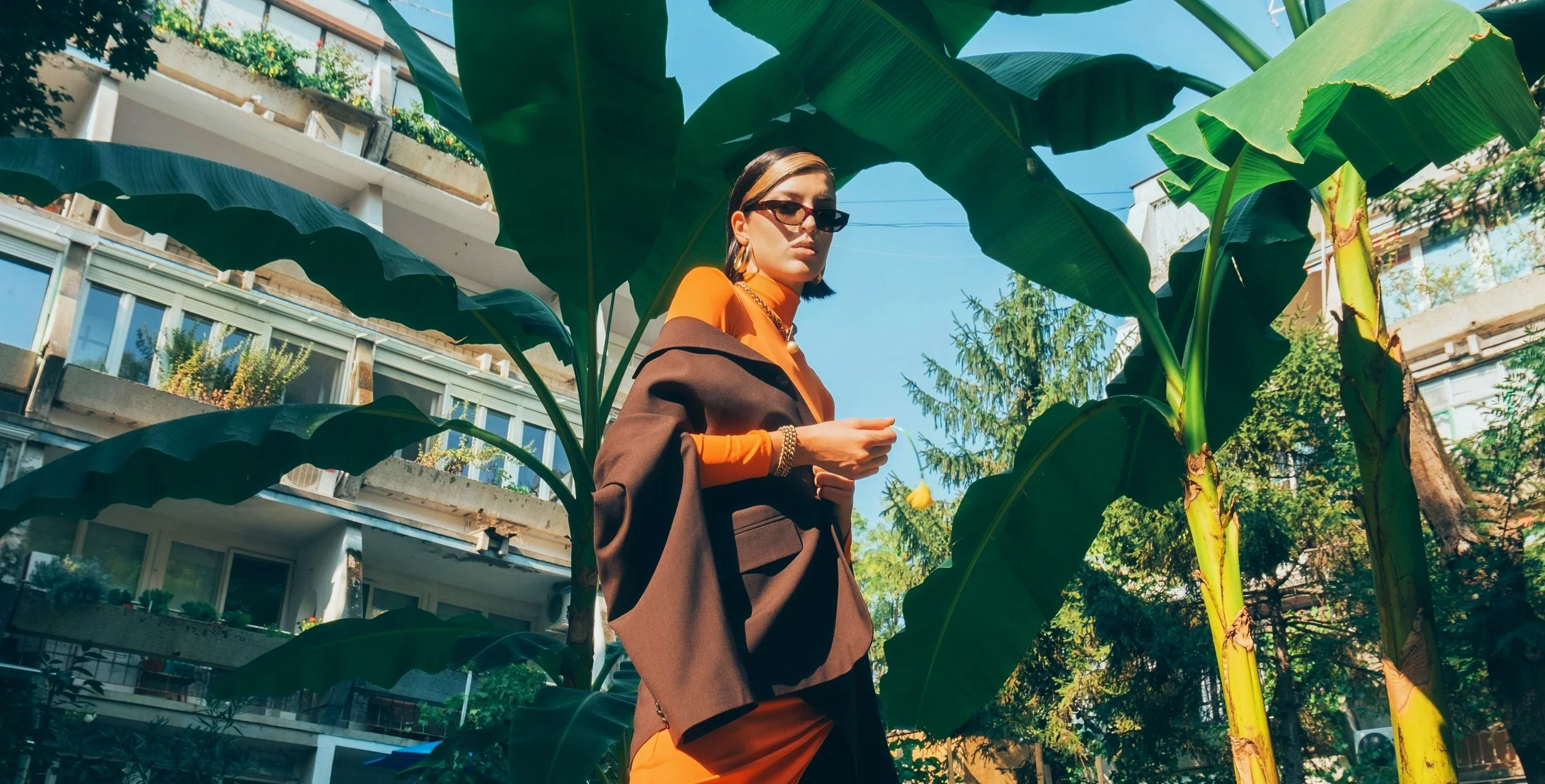 A woman standing amid large green banana leaves, wearing sunglasses, a brown blazer draped over her shoulders, an orange top, and accessories, with a multi-story apartment building in the background under a clear blue sky.