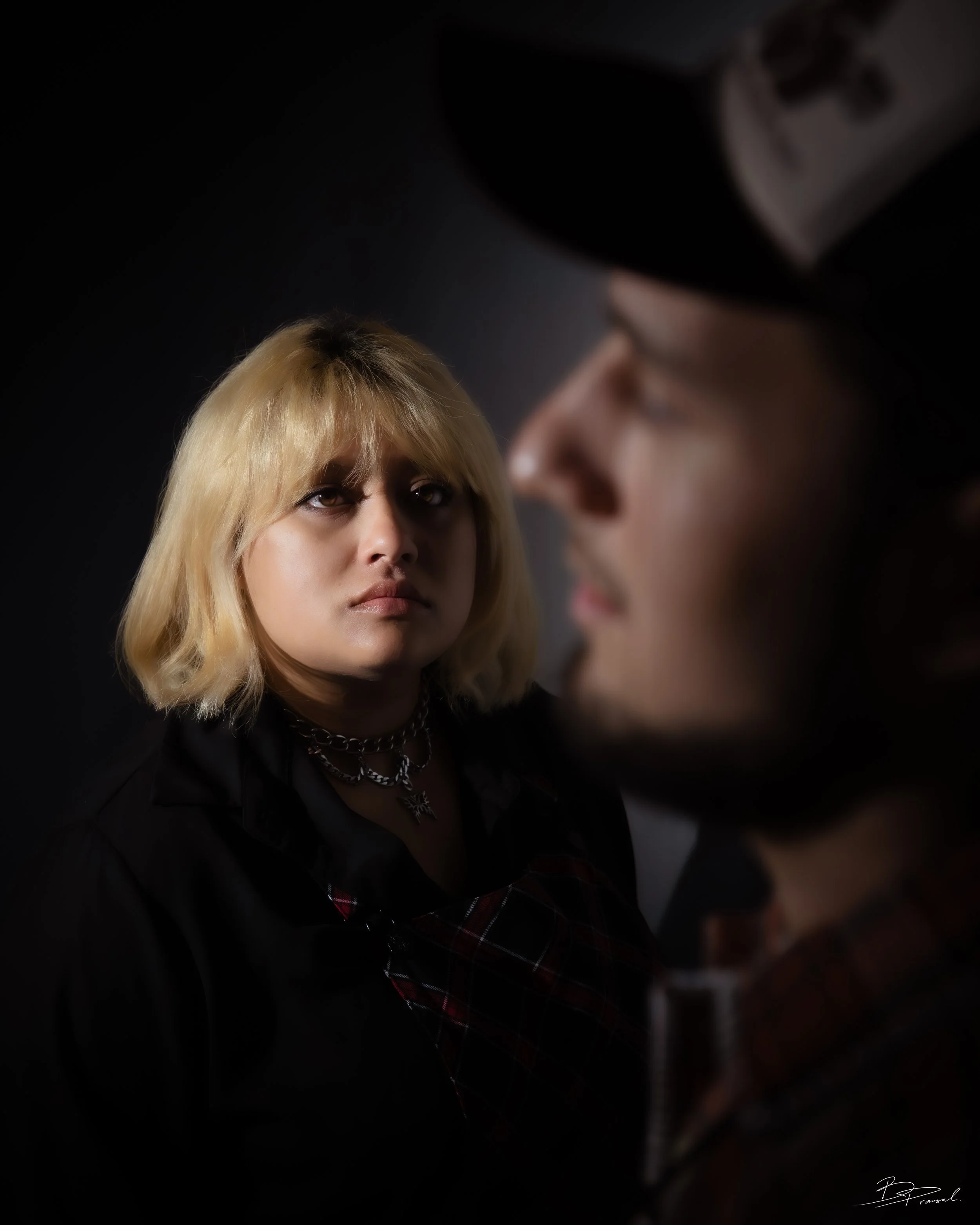 A young woman with platinum blonde hair and dark eye makeup looking intensely at a person wearing a black cap and glasses. The background is dark, highlighting their faces.