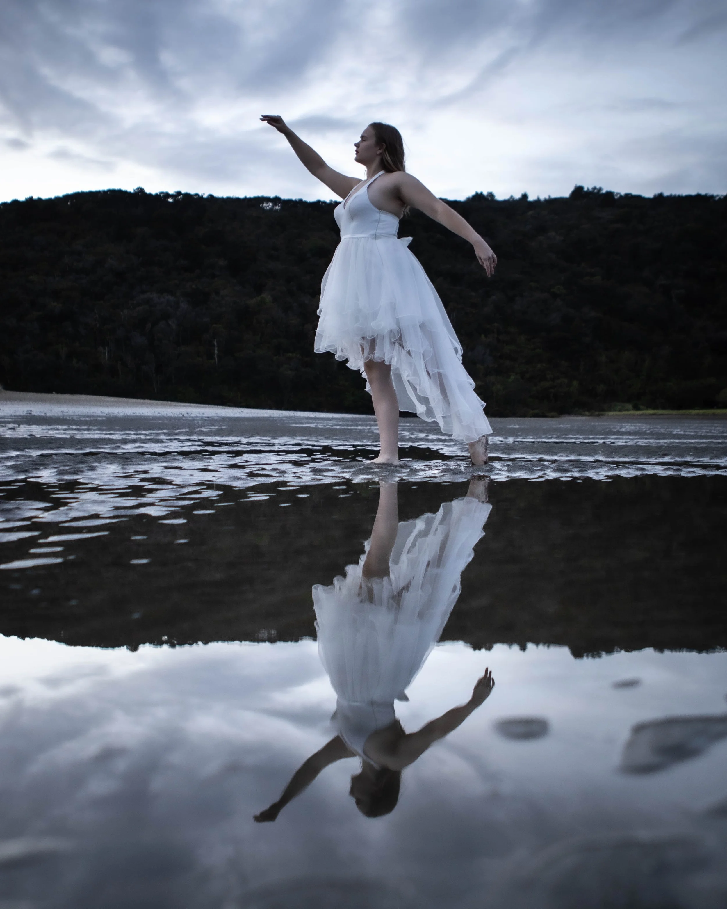 A woman in a white dress standing in shallow water, with her reflection visible beneath her, against a backdrop of hills and a cloudy sky.