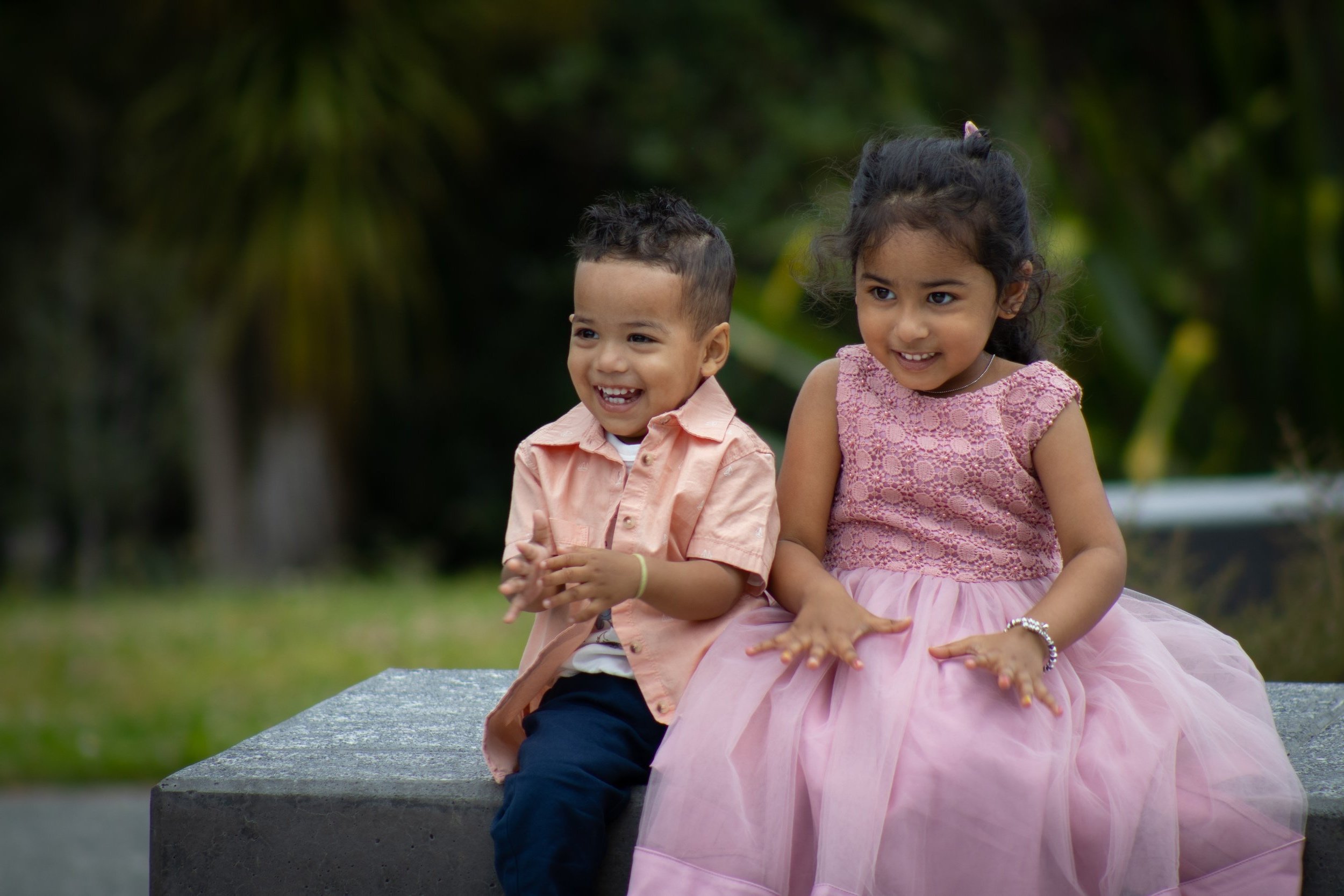 Two young children, a boy and a girl, sitting on a stone bench outdoors, smiling and looking happy. The boy is wearing a light pink shirt and dark pants, and the girl is wearing a pink dress with a lace top.
