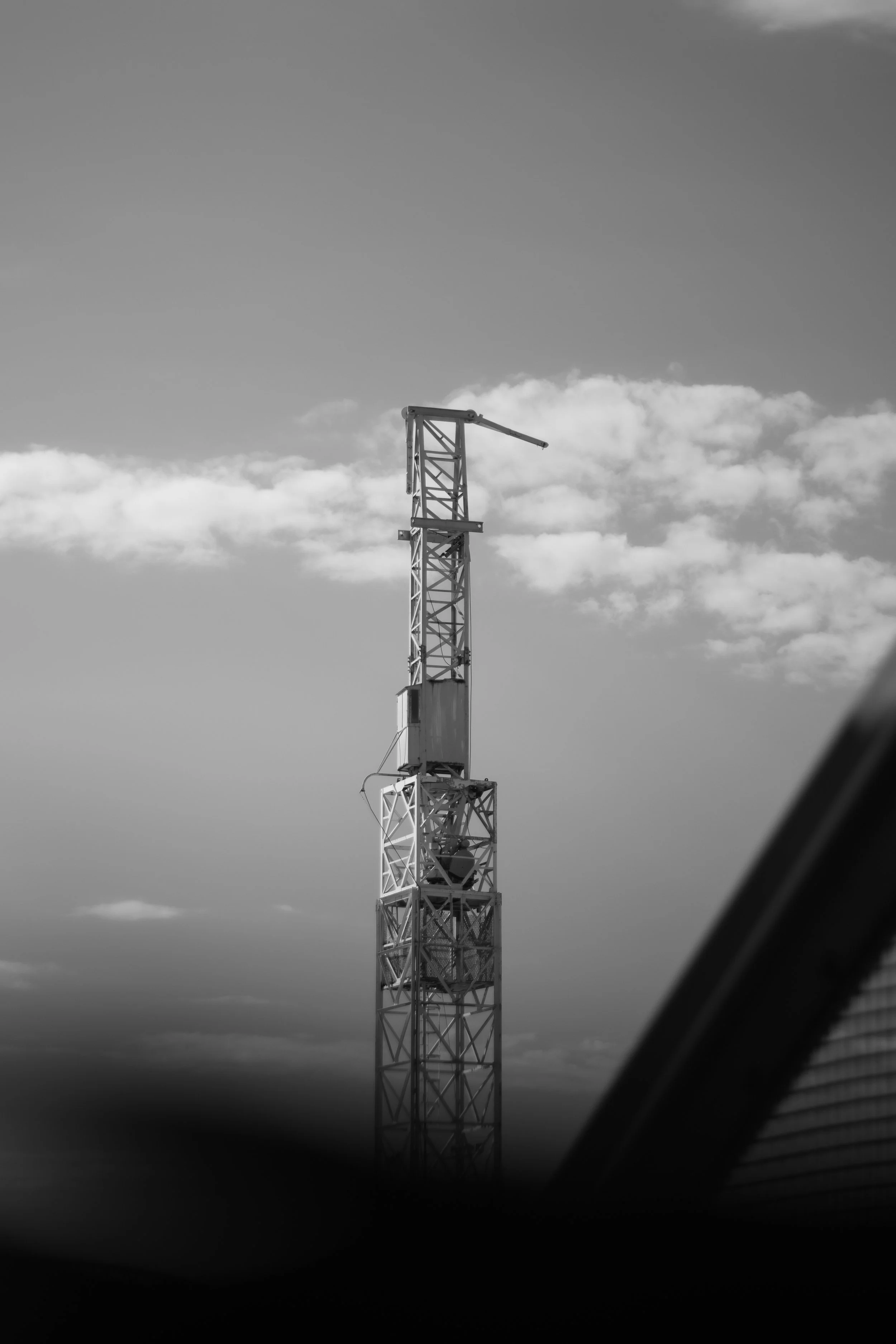 Black and white photo of a tall construction crane against a cloudy sky, with part of a vehicle or structure in the foreground.