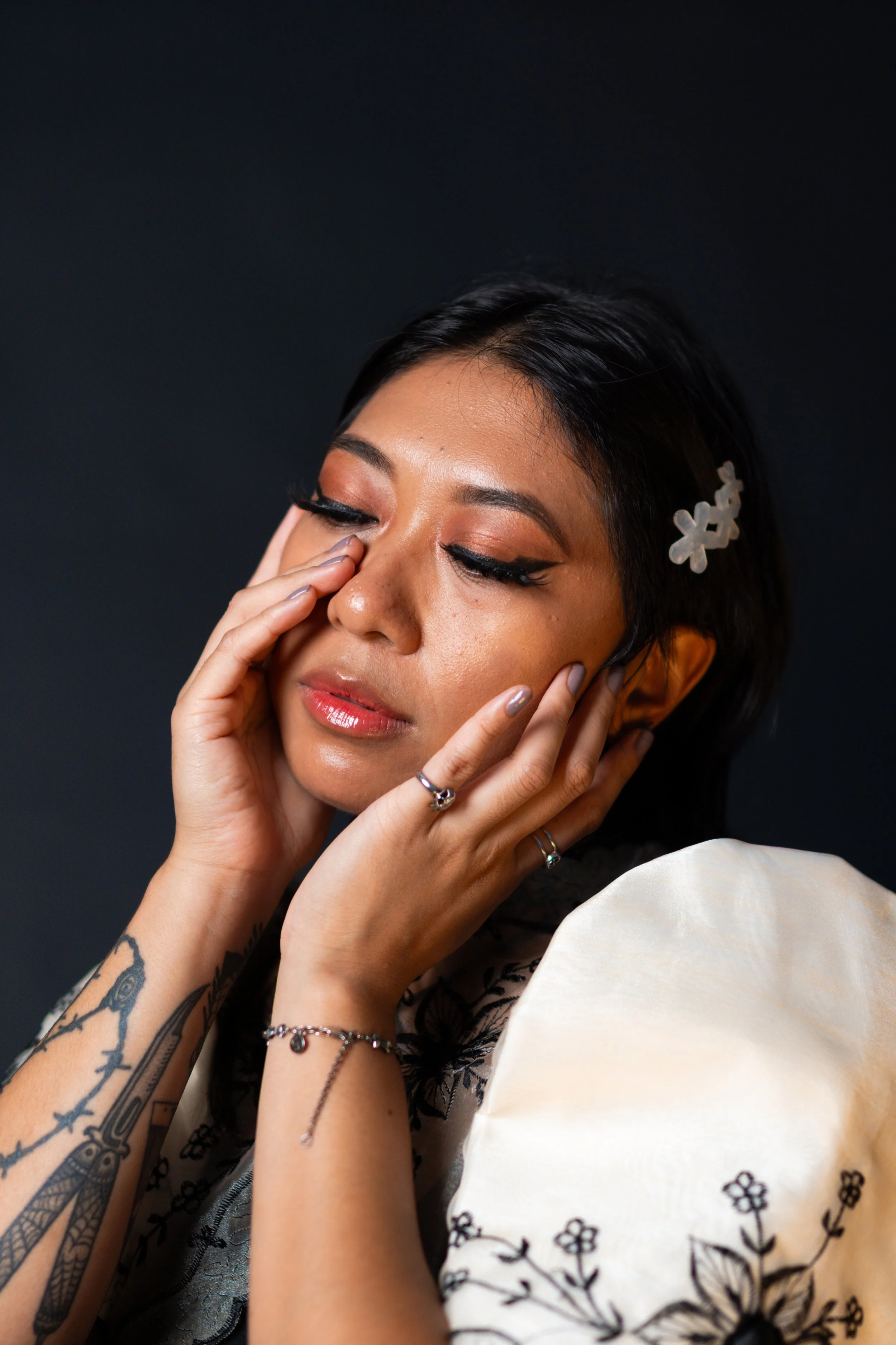 A woman with dark hair, tattoos, and makeup touches her face with her hands, wearing jewelry and a floral-patterned cream-colored blouse, against a dark background.