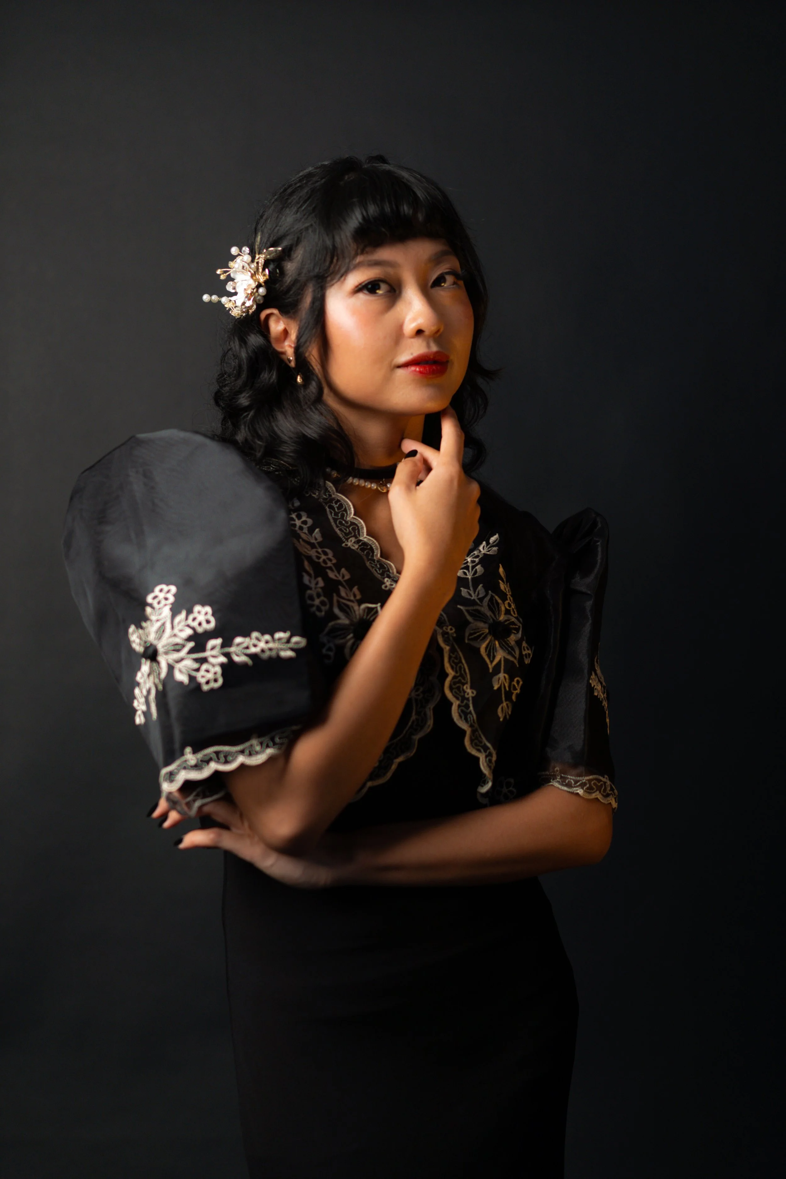 A woman with black hair, wearing a black dress with floral embroidery and puffed sleeves, posing thoughtfully against a dark background.