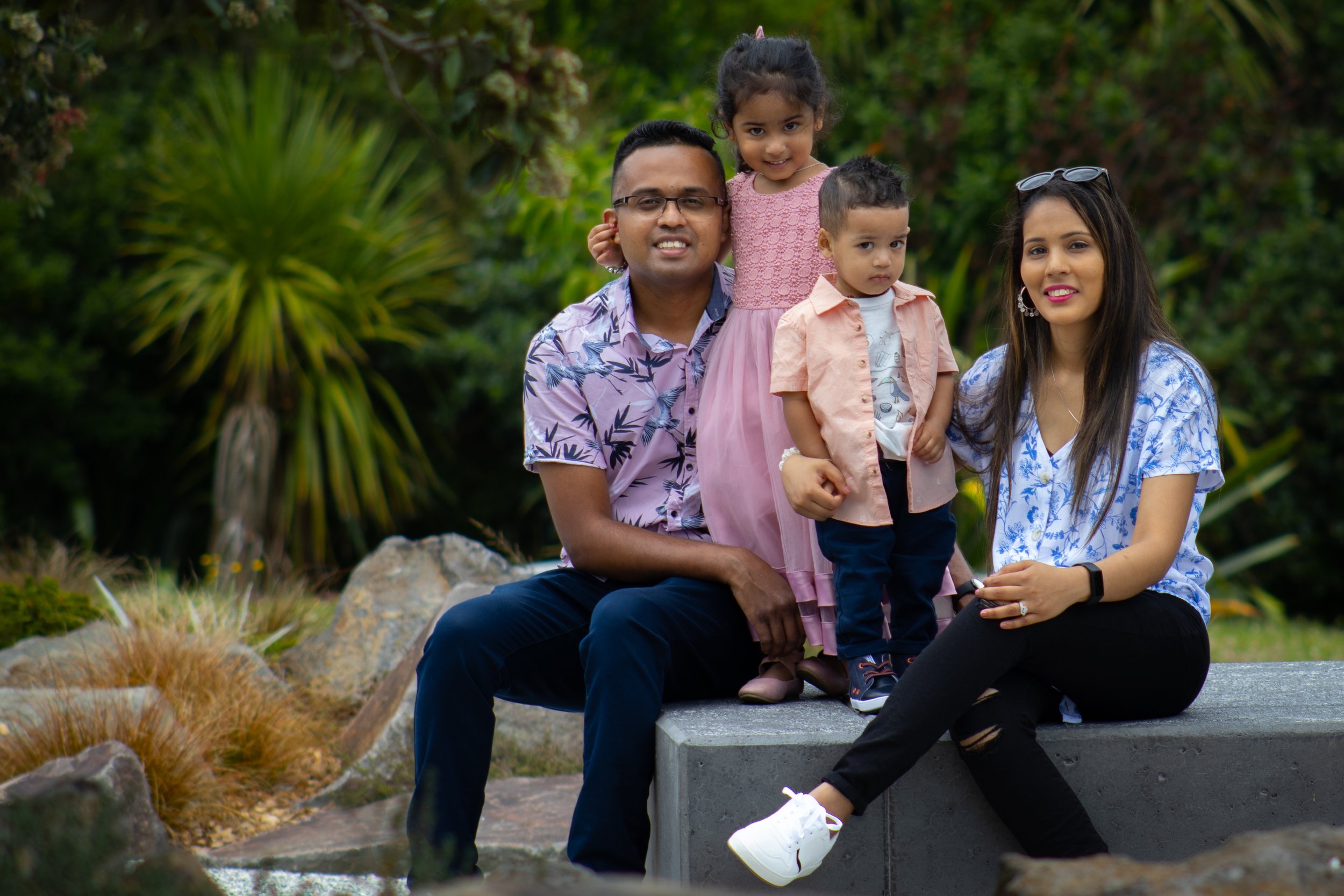 A family of four posing outdoors in a park, sitting and standing on a stone ledge, with greenery and plants in the background.
