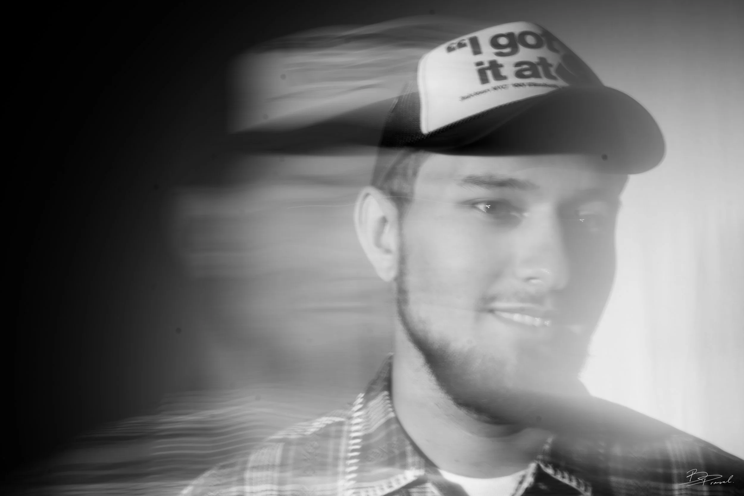 Black and white photo of a young man wearing a baseball cap with text on the front, looking at the camera with a slight smile, with motion blur on one side.