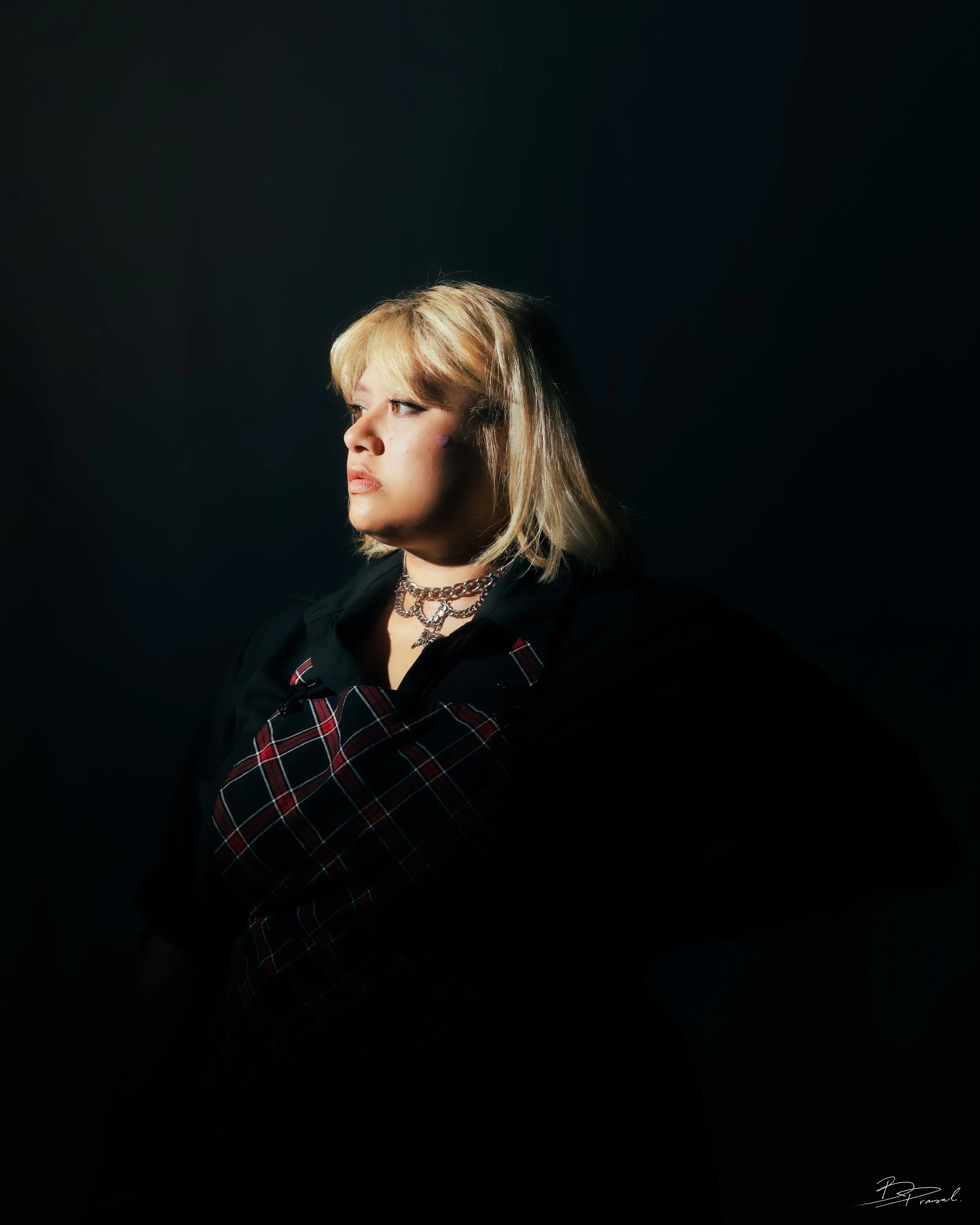 A woman with blonde hair wearing a black shirt, plaid suspenders, and layered necklaces, gazing to the left against a dark background.