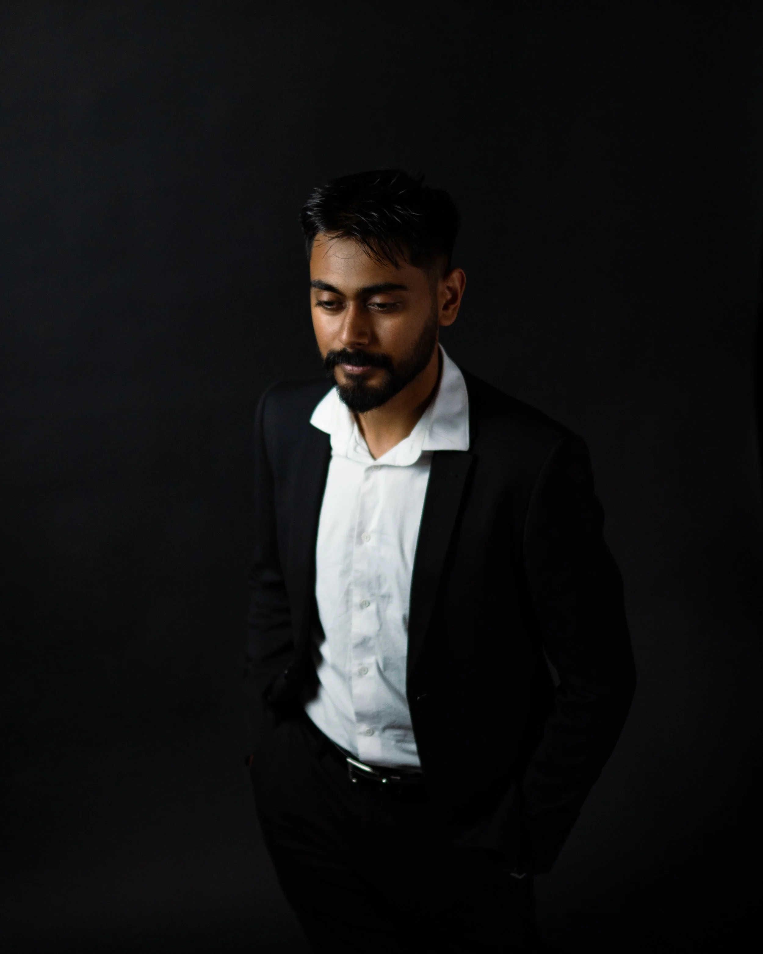 A man in a black suit with a white shirt, standing against a dark background, looking down with his hands in his pockets.