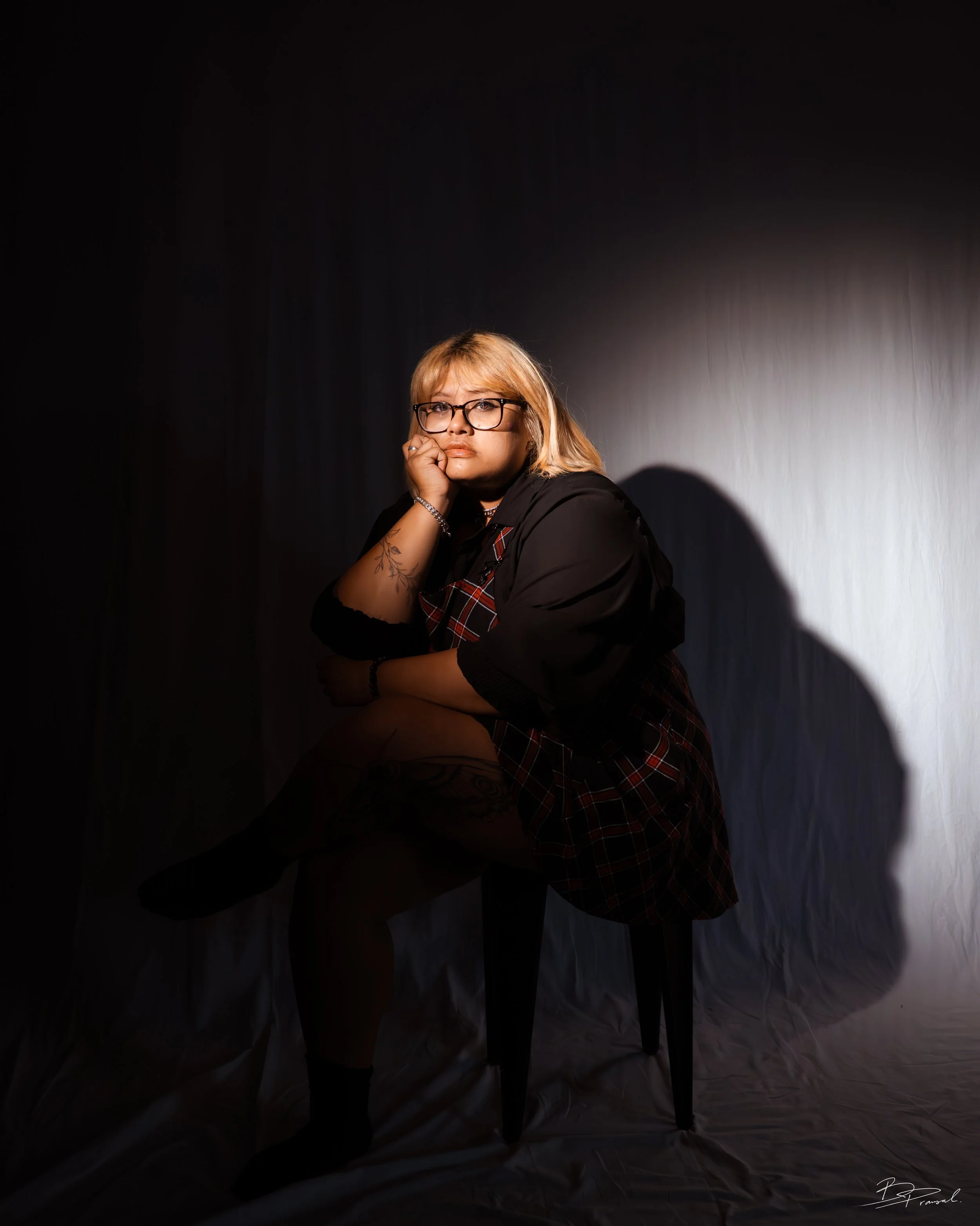 A woman with blonde hair, glasses, and tattoos sitting on a chair against a dark background, with her head resting on her hand and looking contemplative.
