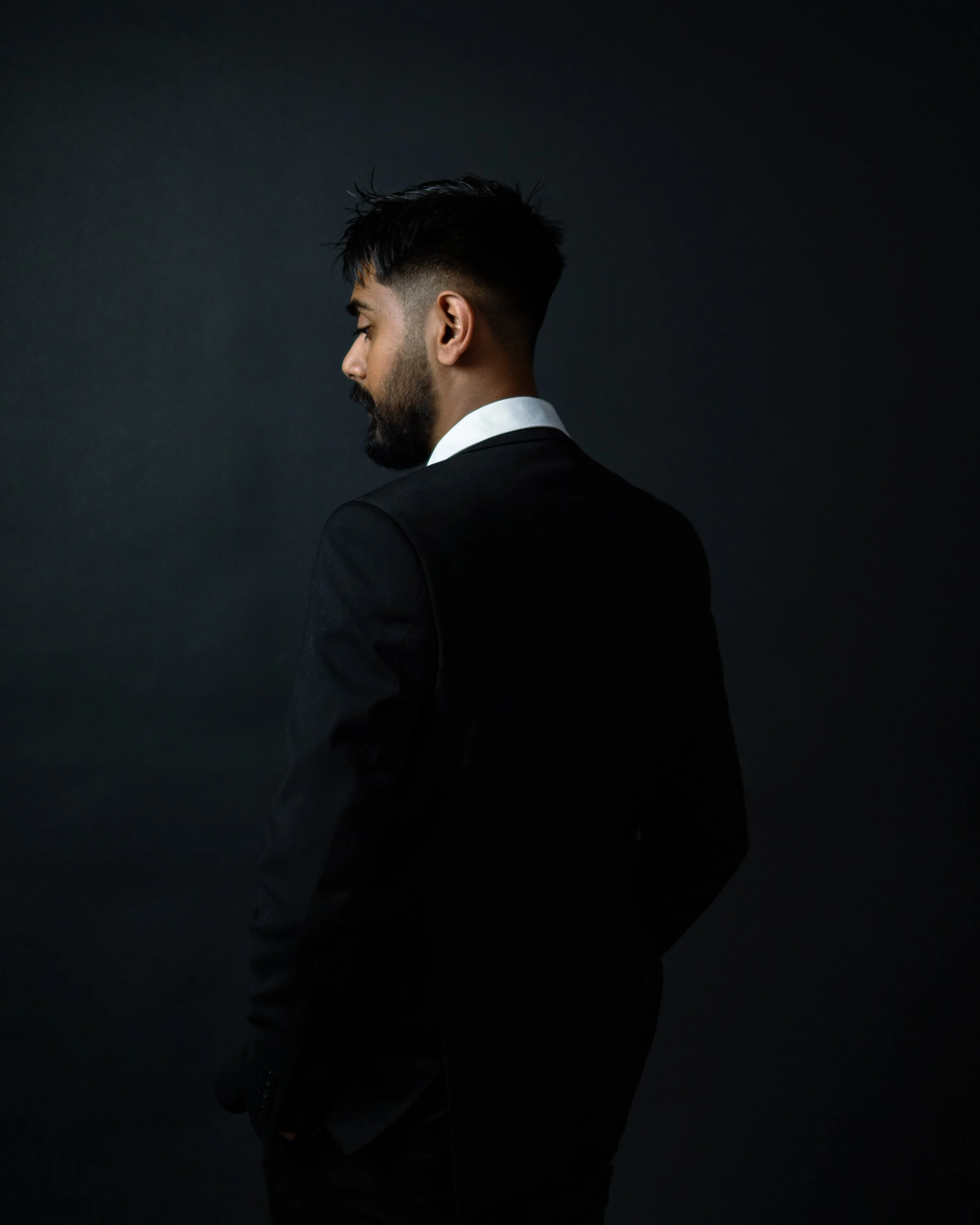 Side profile of a man with a beard in a black suit and white shirt, standing against a dark background.