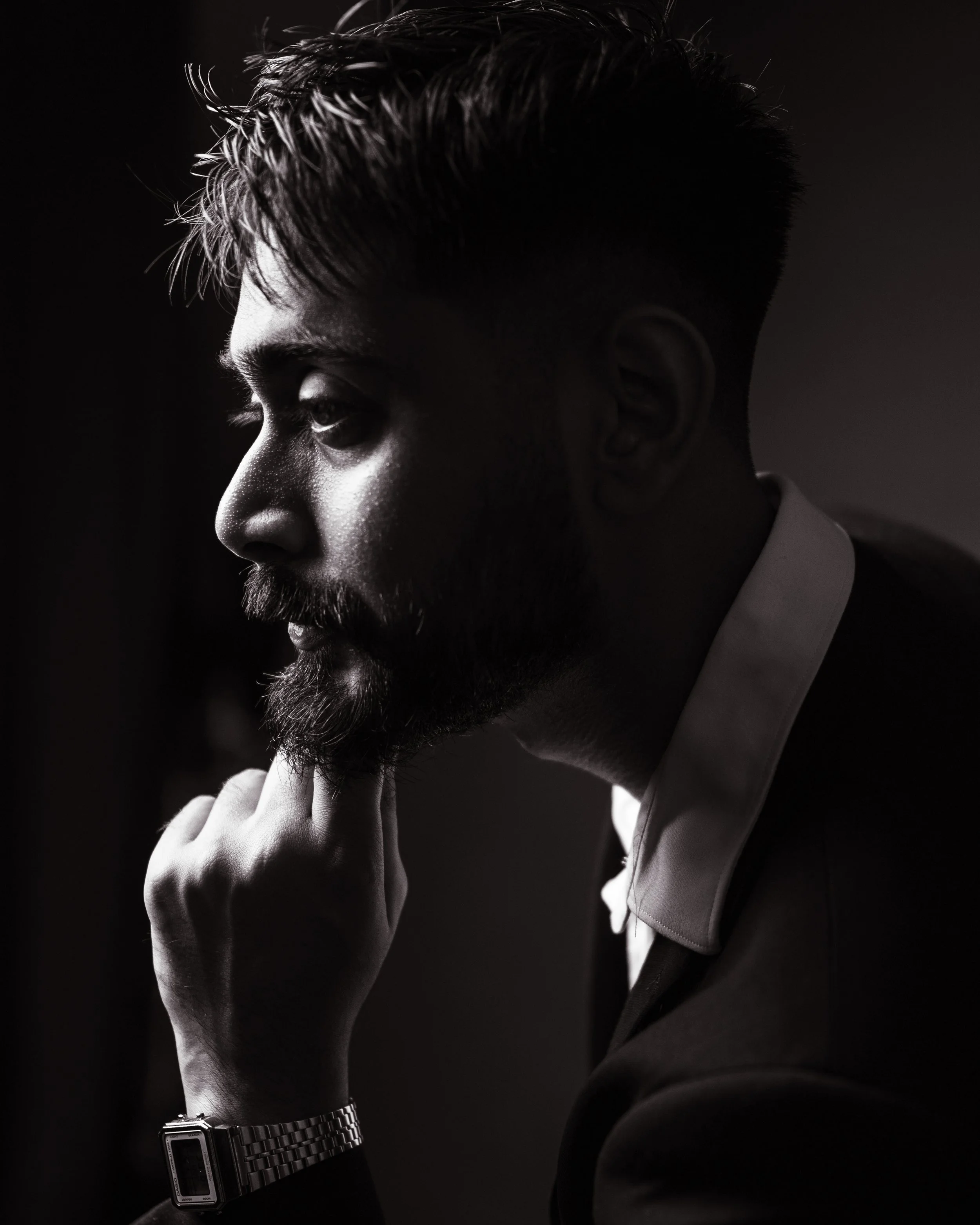 Black-and-white side profile portrait of a man with a beard, wearing a collared shirt and a wristwatch, resting his chin on his hand in deep thought.