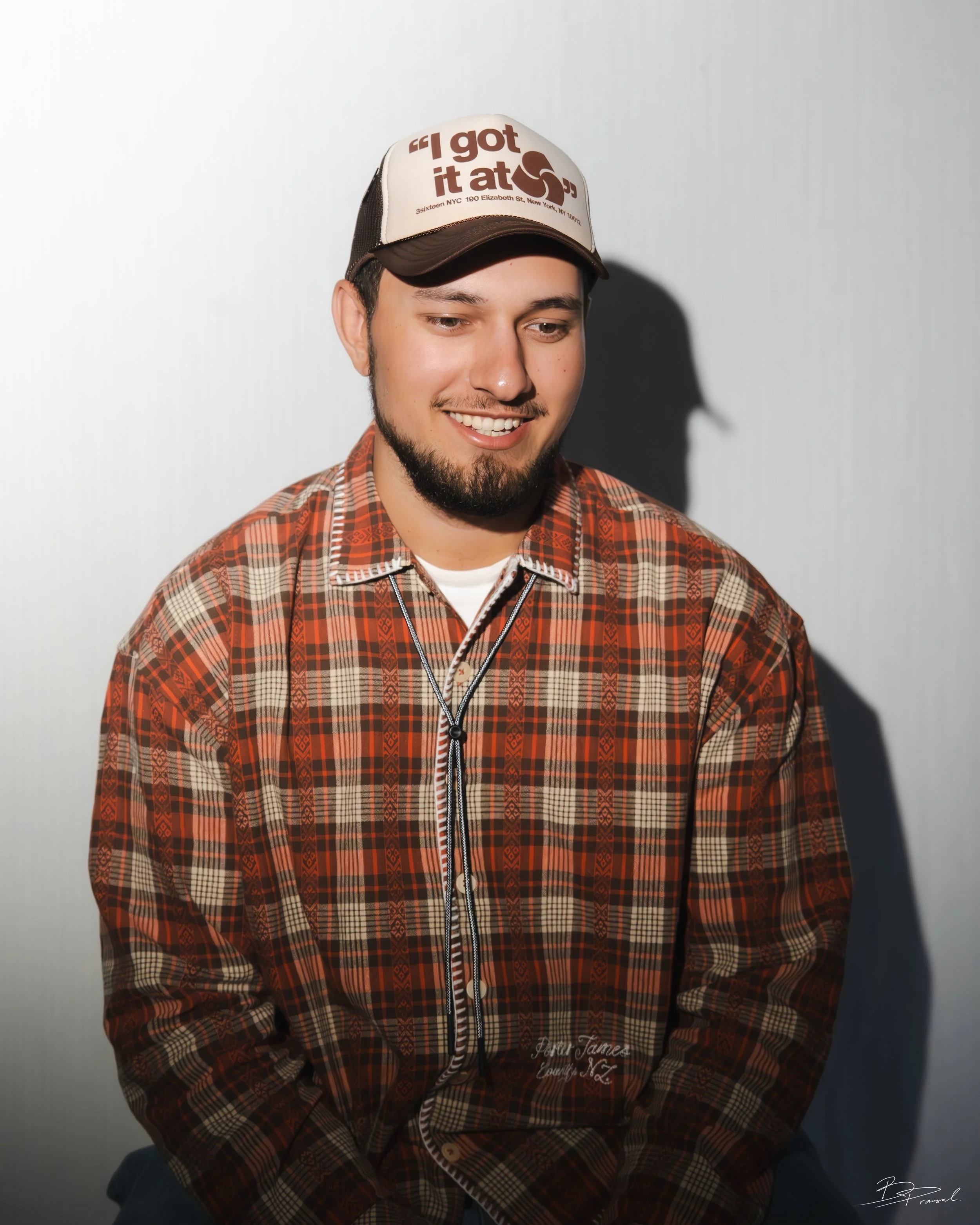 A young man with a beard and mustache wearing a plaid shirt and a baseball cap, smiling and looking down, with a shadow cast on a light background behind him.