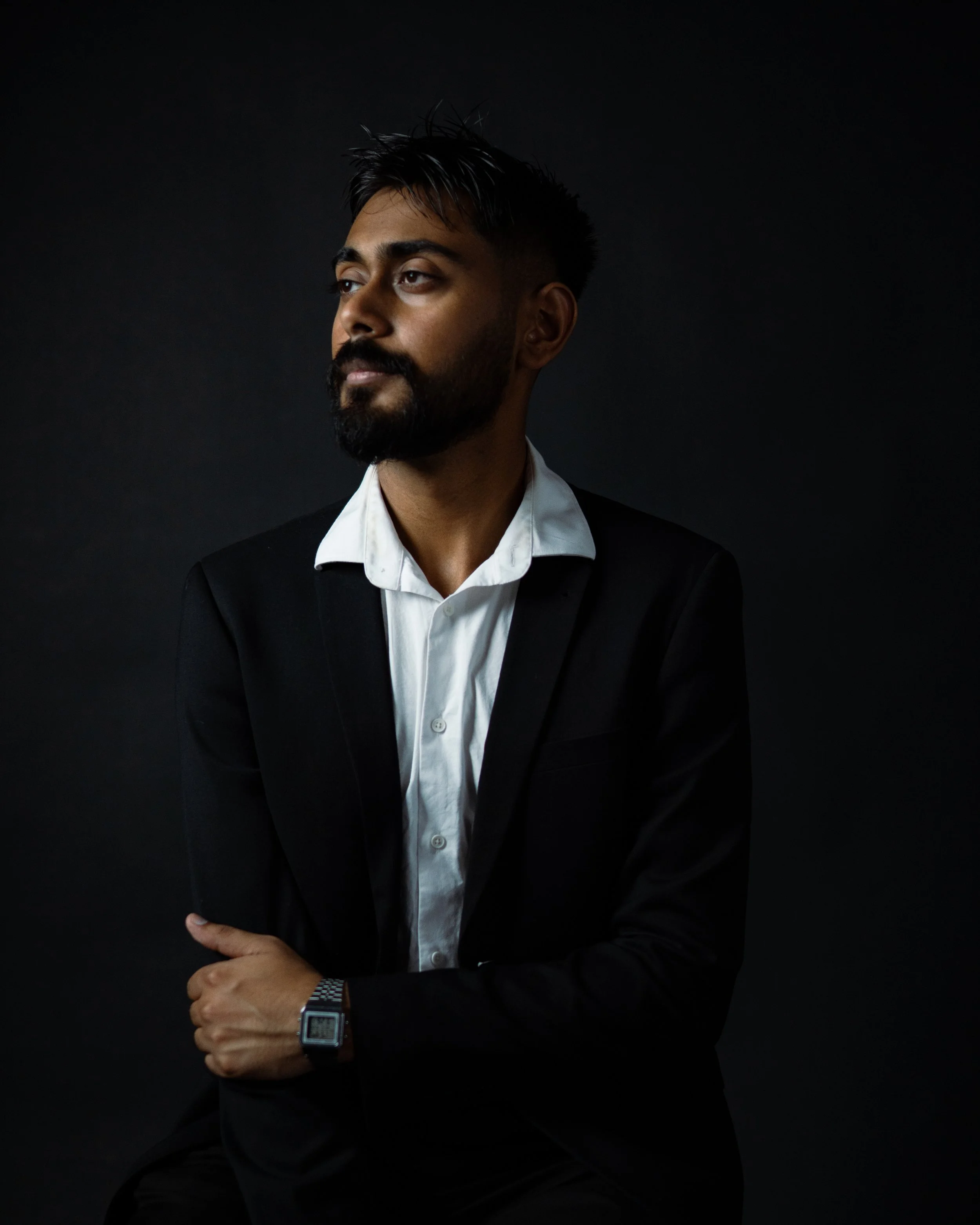A man with a beard and styled hair wearing a black suit and white shirt, sitting with his arm crossed, against a black background.