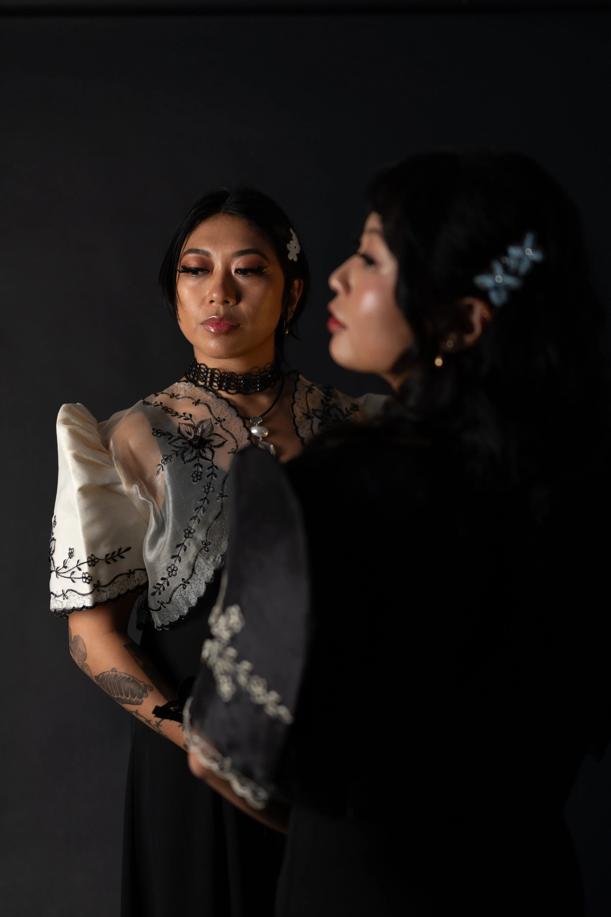 Two women with black hair and tattoos on their arms, wearing elegant clothing and jewelry, are facing each other against a black background.
