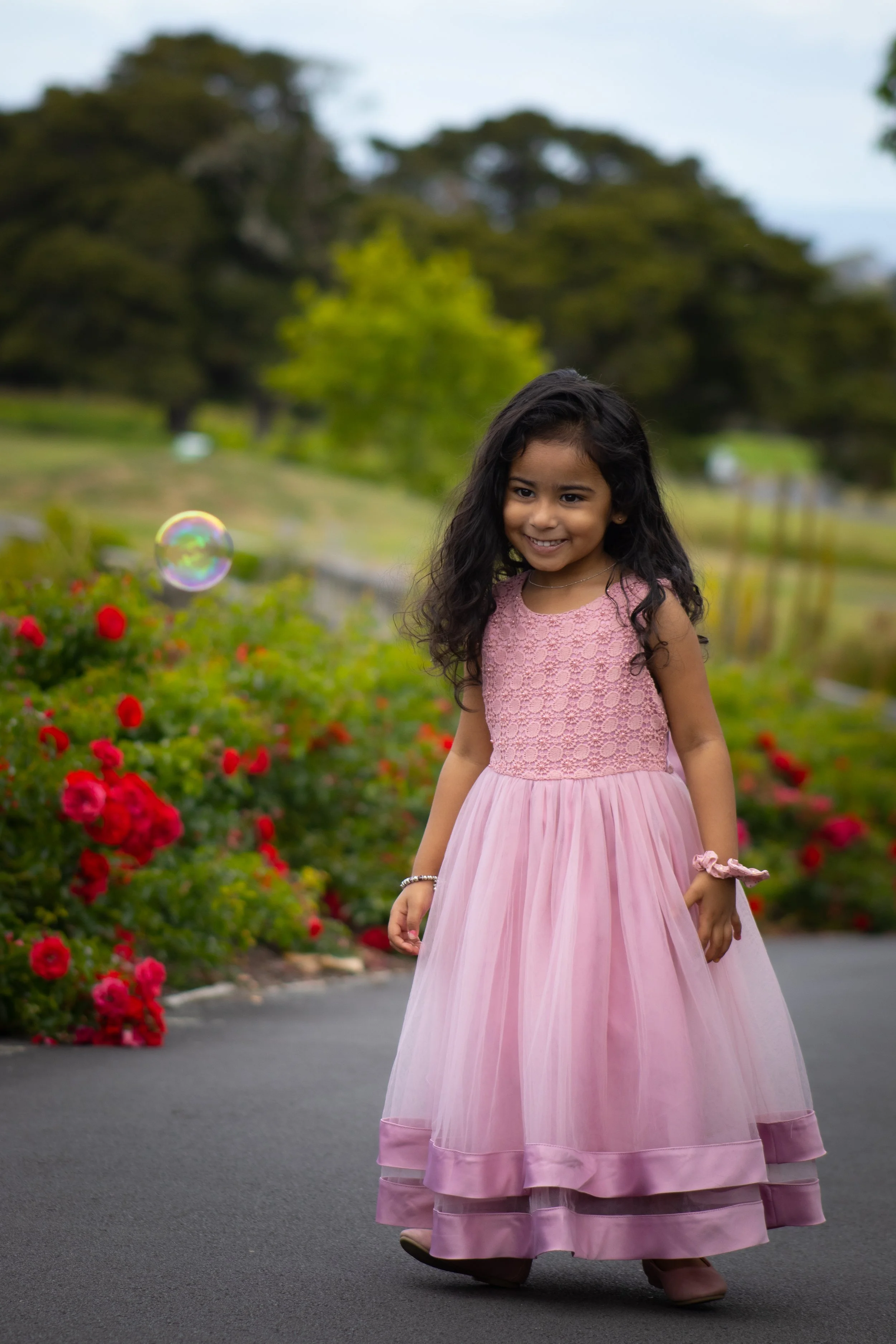 A young girl with long dark hair wearing a pink dress standing on a paved path in a garden with red flowers and trees, smiling at the camera with a colorful soap bubble floating nearby.