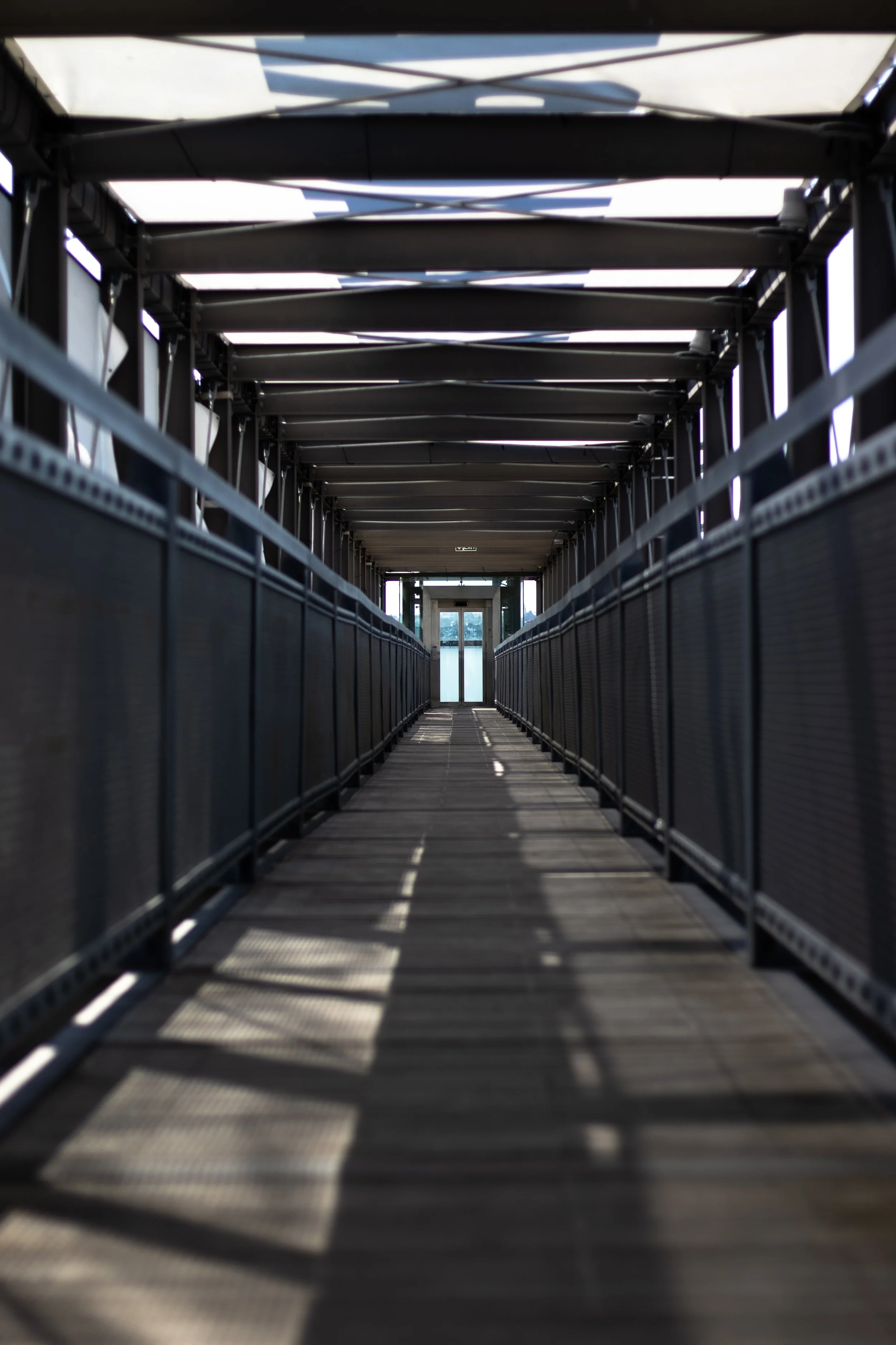 An empty pedestrian bridge with shadow patterns on the wooden walkway, metal railings, and a set of glass doors at the end.