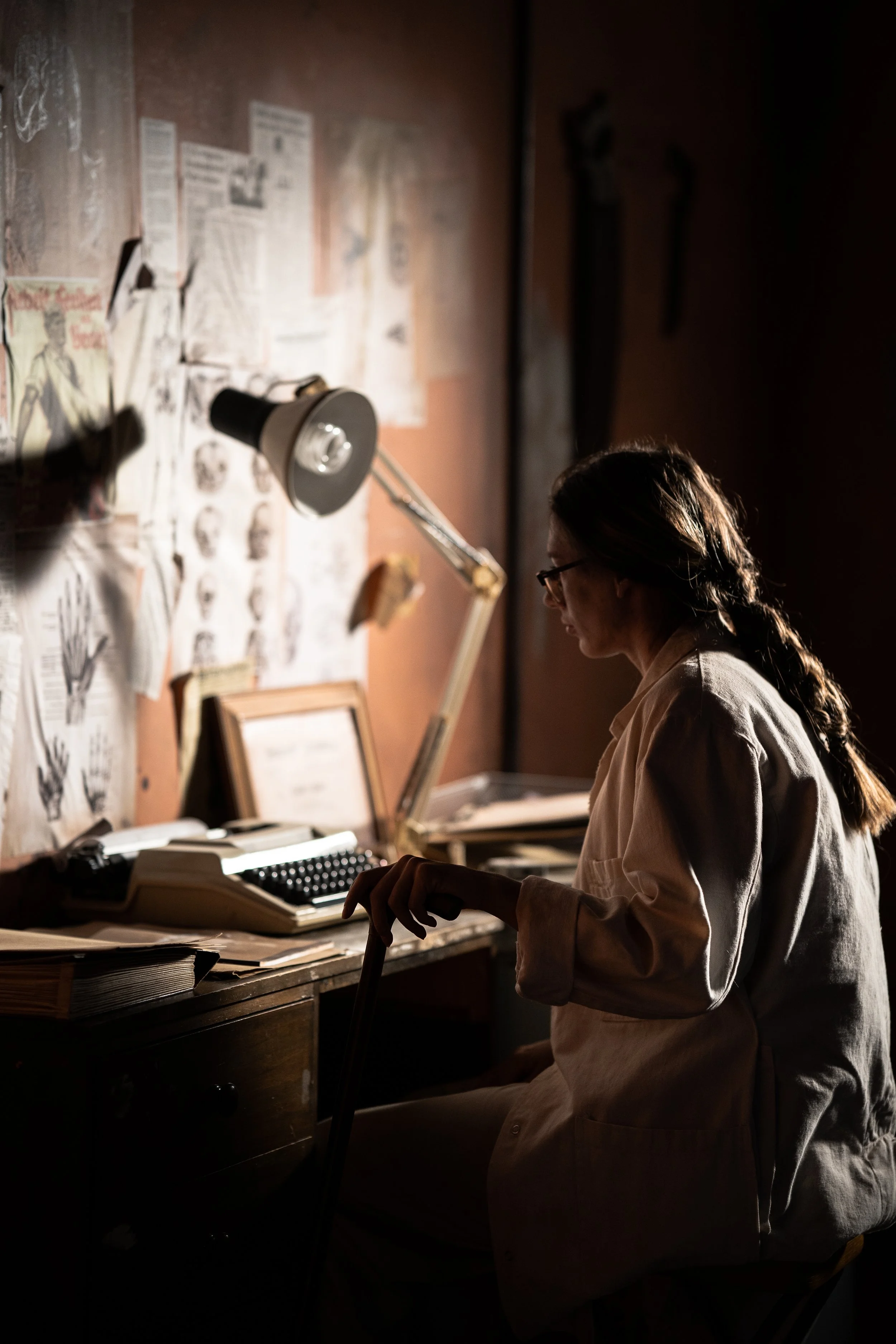 A woman in glasses and a white lab coat sitting at a cluttered desk, working with papers and a typewriter in a dimly lit room with a corkboard wall covered in notes and sketches.