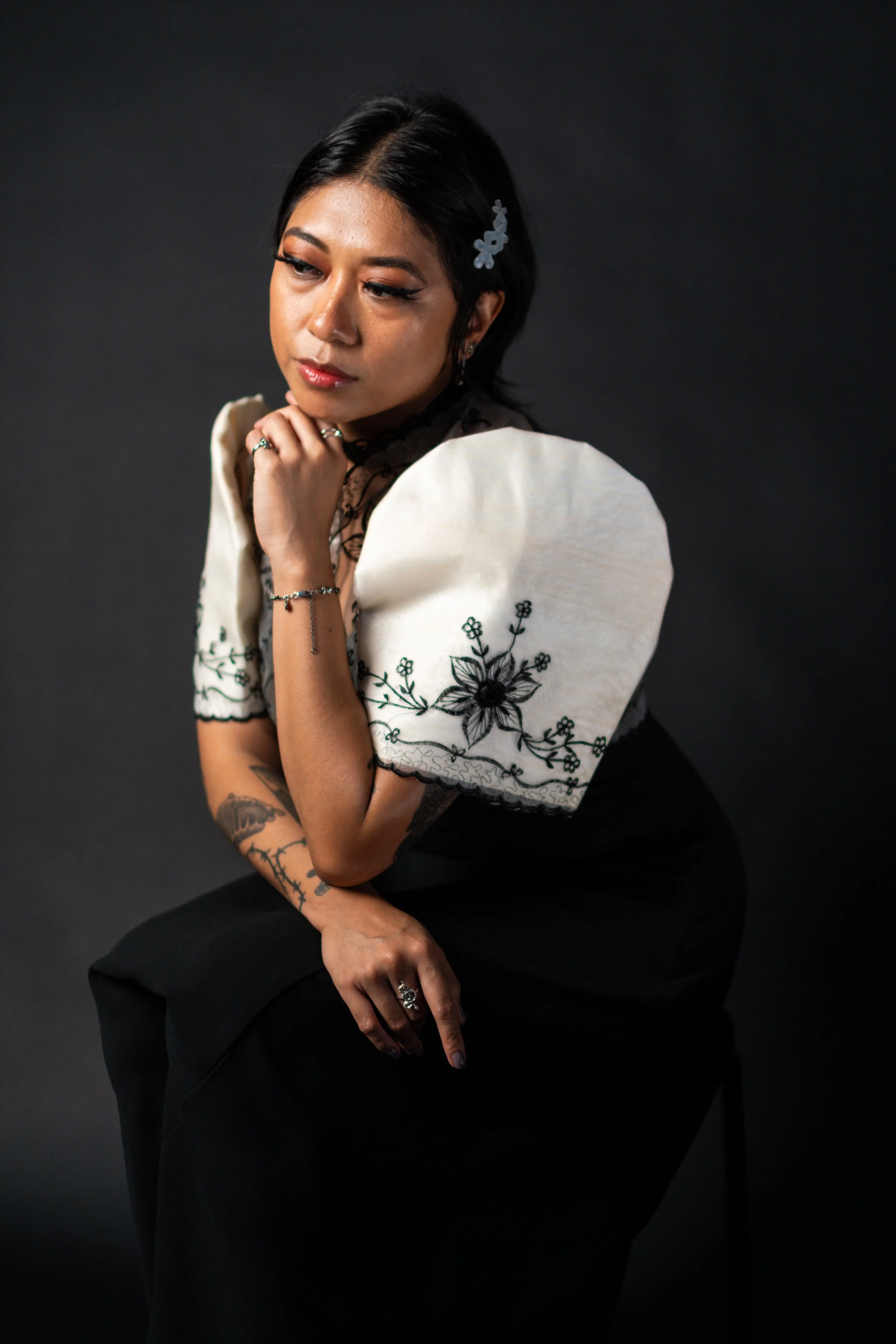 A woman with black hair, tattoos on her arms, and jewelry, sitting against a dark background, wearing a cream-colored embroidered top and black pants.