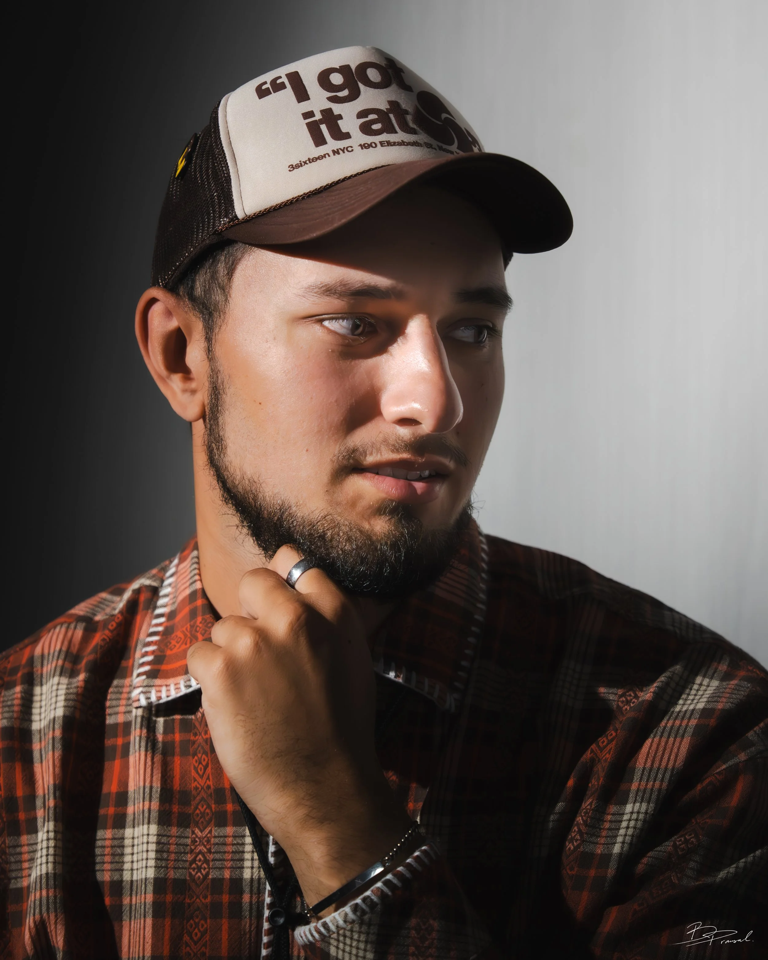Portrait of a young man wearing a trucker hat and a plaid shirt, with his hand near his chin, looking to the side in a studio setting.