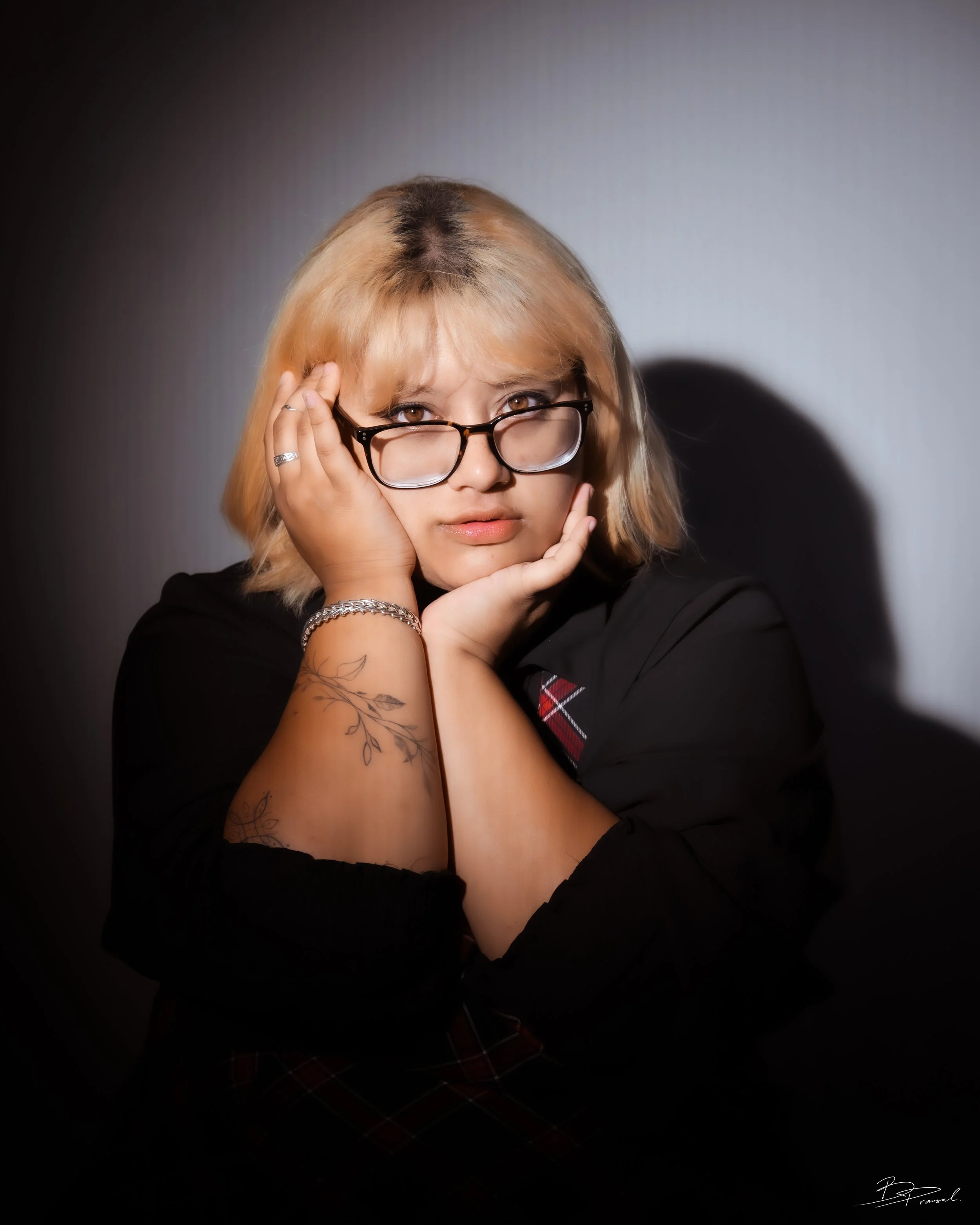 A woman with blonde hair, glasses, and tattoos on her arm, resting her face in her hands, looking directly at the camera with a thoughtful expression, against a plain background.