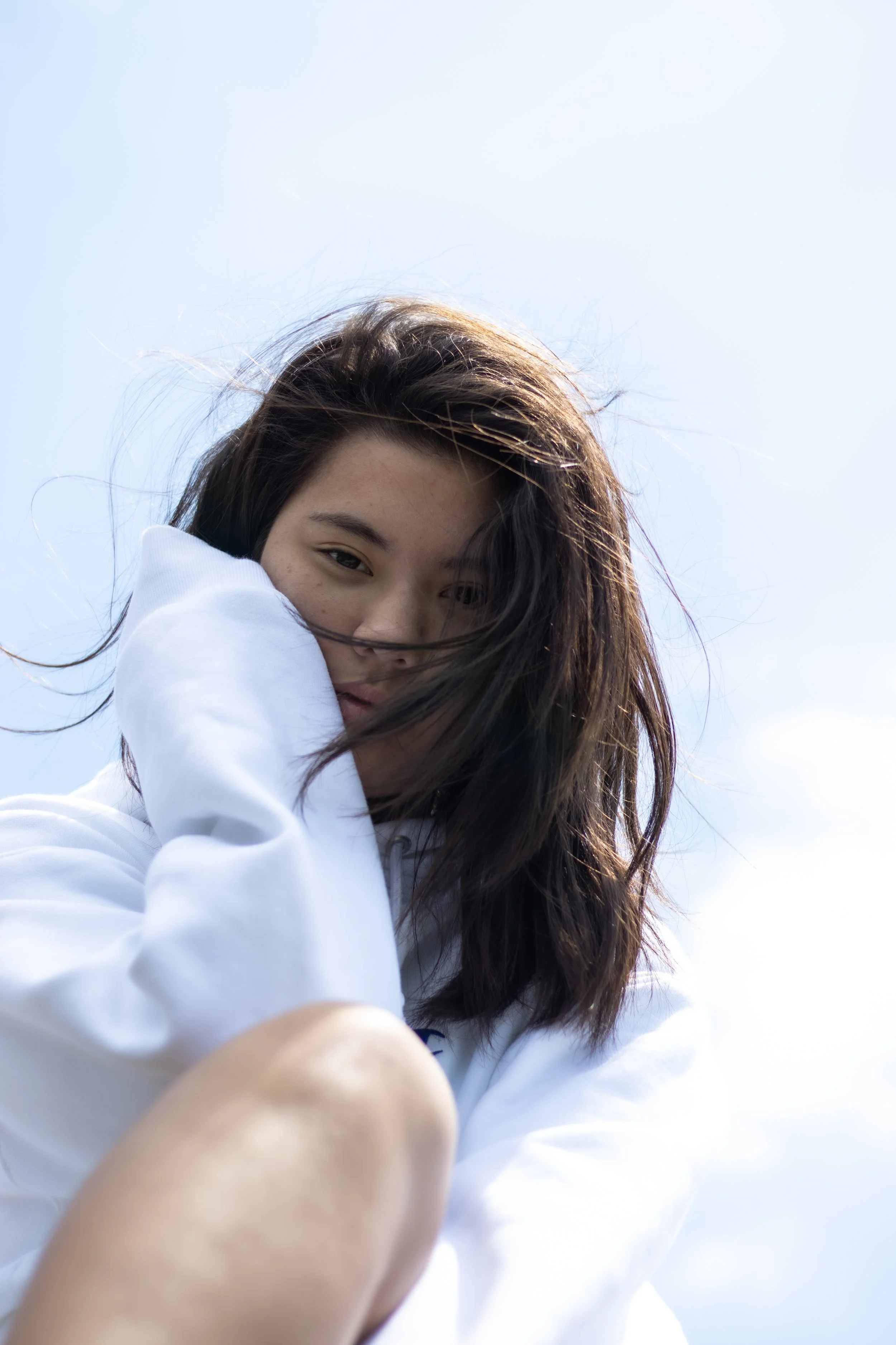Young woman with shoulder-length brown hair, wearing a white hoodie, sitting outdoors against a partly cloudy sky.