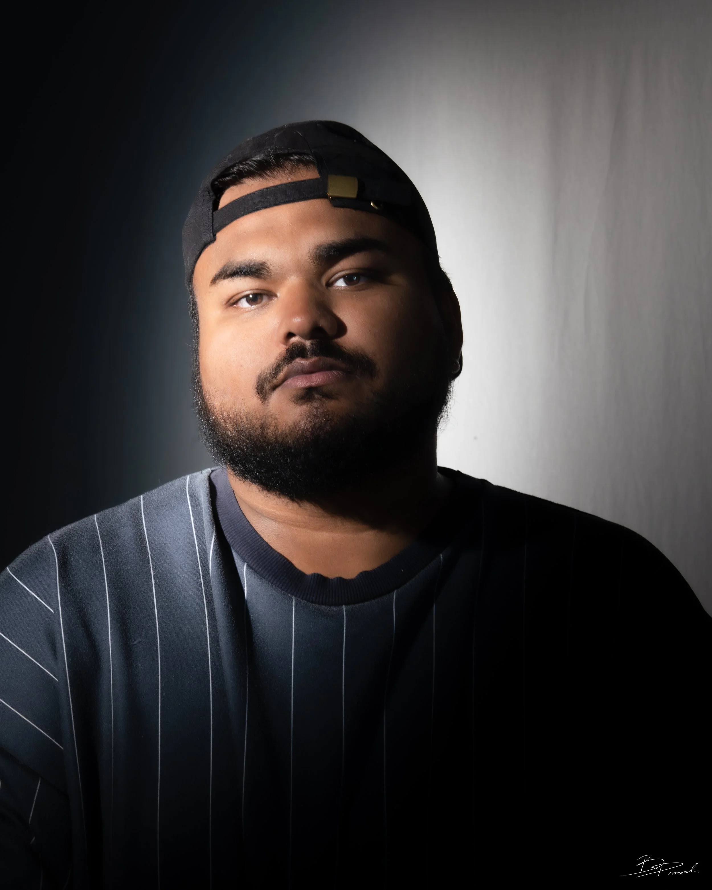 Portrait of a young man with a beard wearing a black baseball cap backwards and a black striped shirt, standing against a plain background with dramatic lighting from the side.