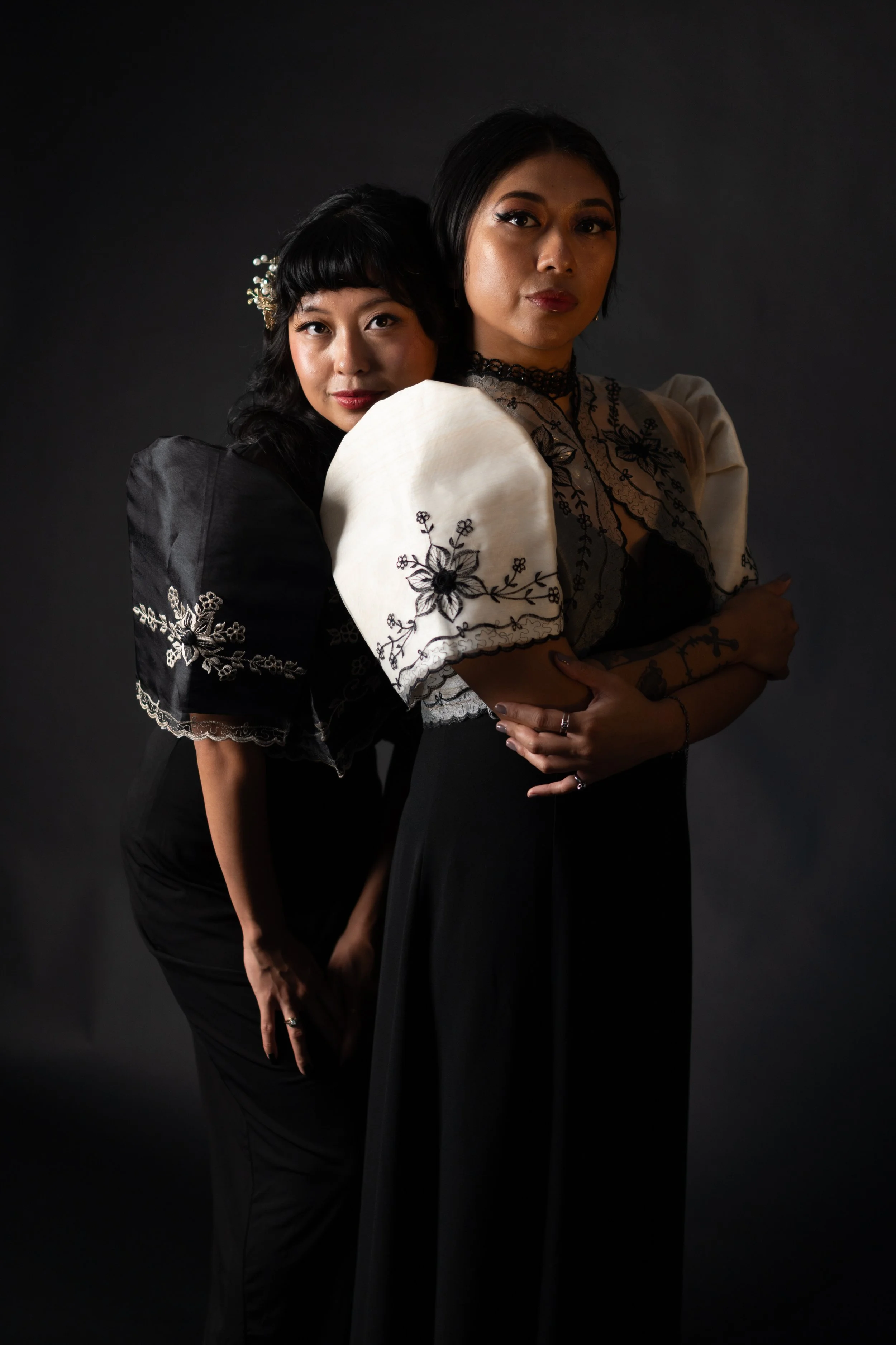 Two women standing close together in a dark studio background, both wearing black and white embroidered clothing, with one woman slightly behind the other, both looking at the camera.