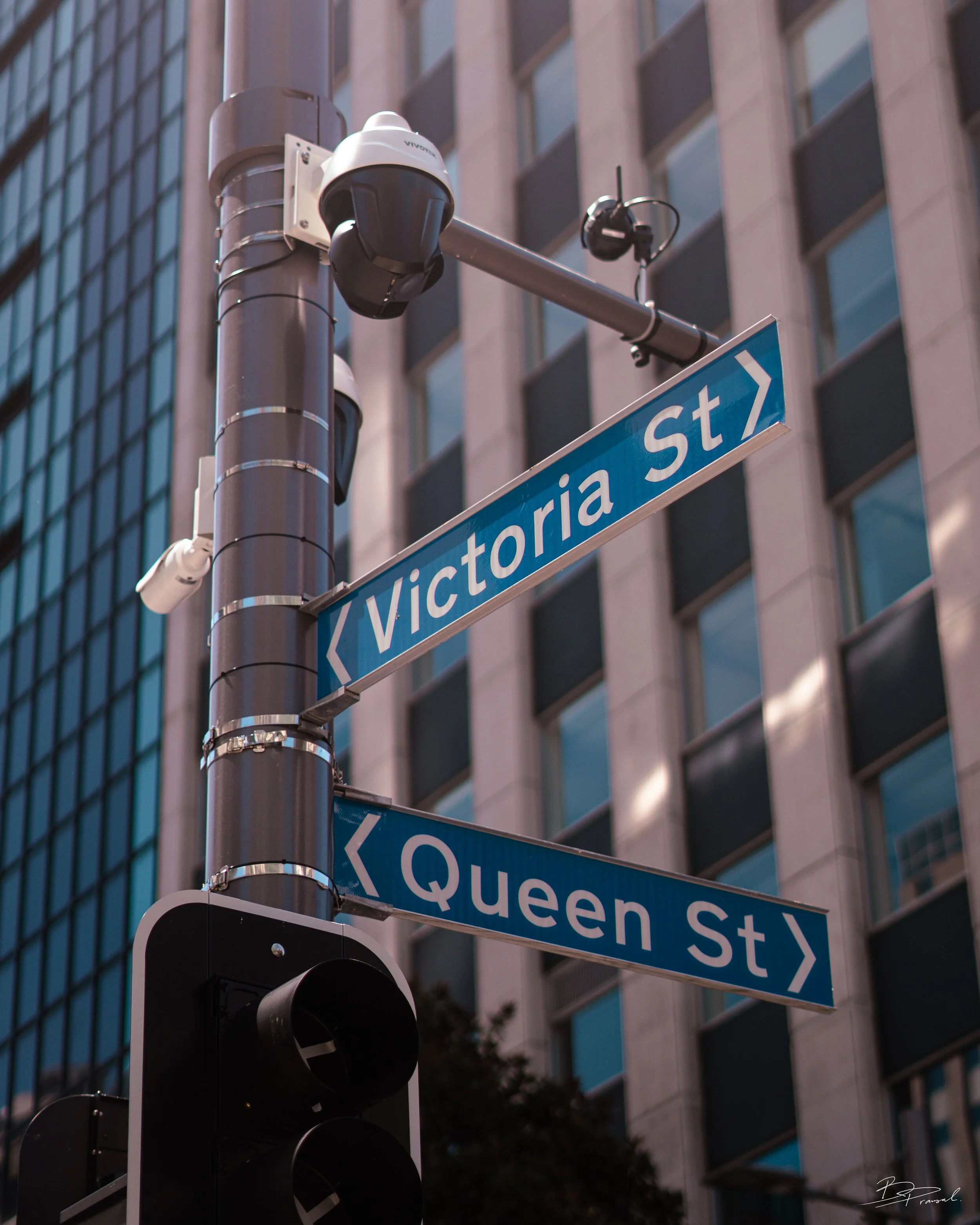 Street sign showing intersection of Victoria Street and Queen Street in an urban area with a traffic camera and high-rise buildings.