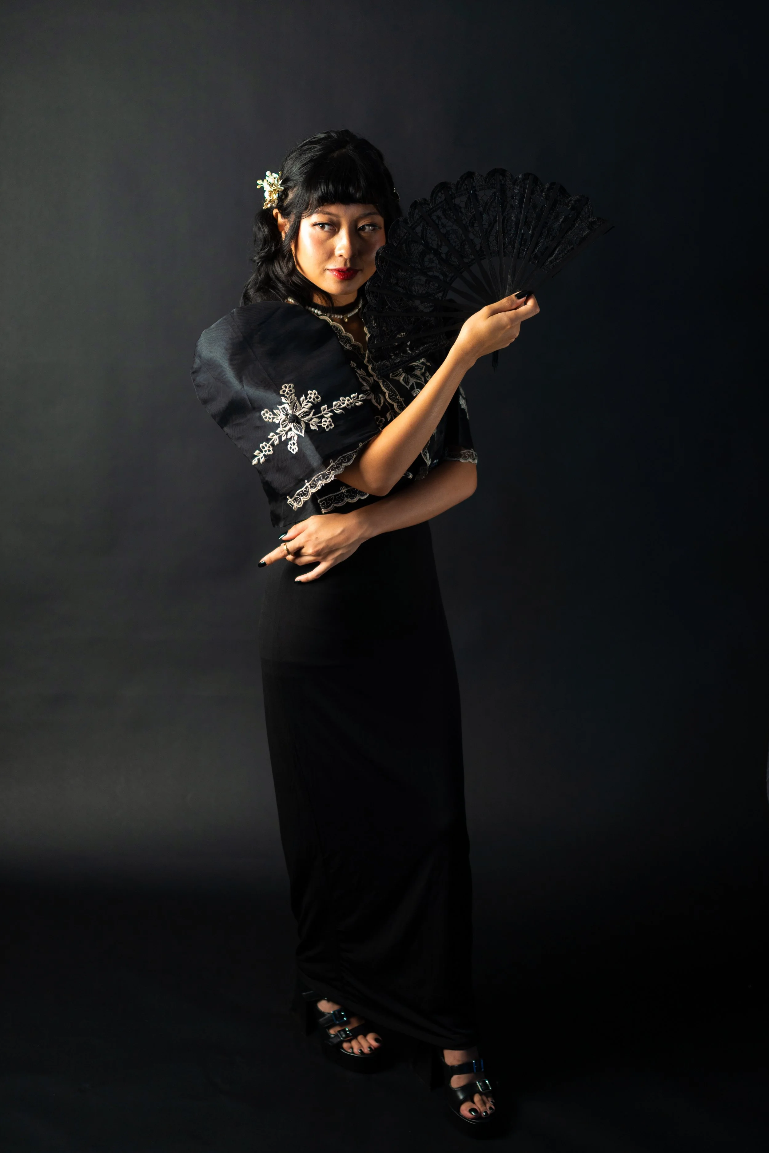 A woman wearing a black dress with embroidered sleeves is holding a black lace fan in front of a dark background.