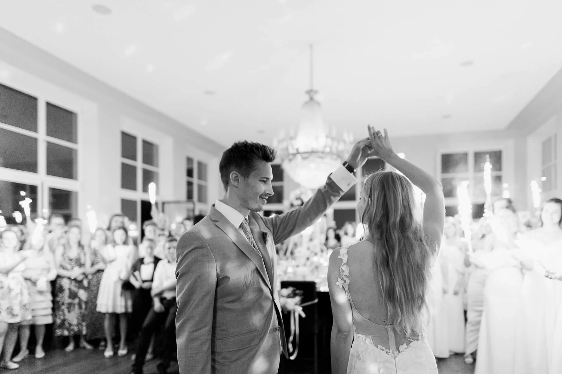Ein Hochzeitspaar tanzt in einem festlich dekorierten Raum, umgeben von Gästen, von denen einige Wunderkerzen in den Händen halten.