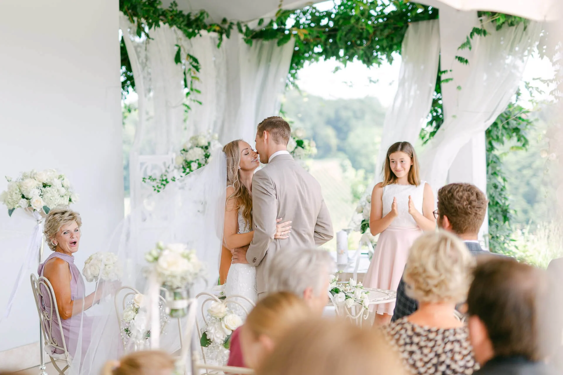 Hochzeitspaar bei Zeremonie, umgeben von Gästen. Braut trägt weißen Schleier und Brautkleid, Bräutigam im Anzug. Dekoration mit Blumen und grünem Blattwerk. Junge Frau klatscht Applaus. Feierliche Stimmung im Freien.