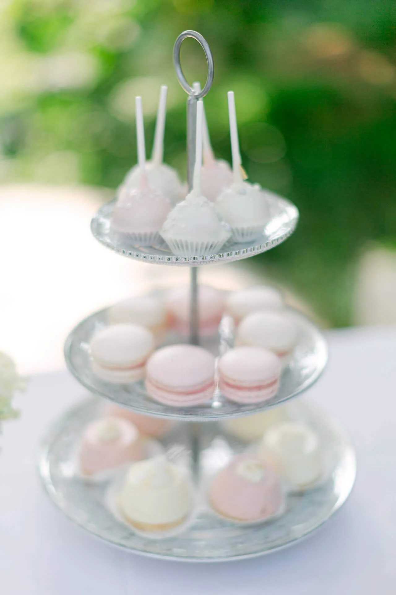 Eine dreistufige Etagere mit weiß-rosa Cake Pops, Macarons und Gebäck, auf einem Tisch mit unscharfem Hintergrund.