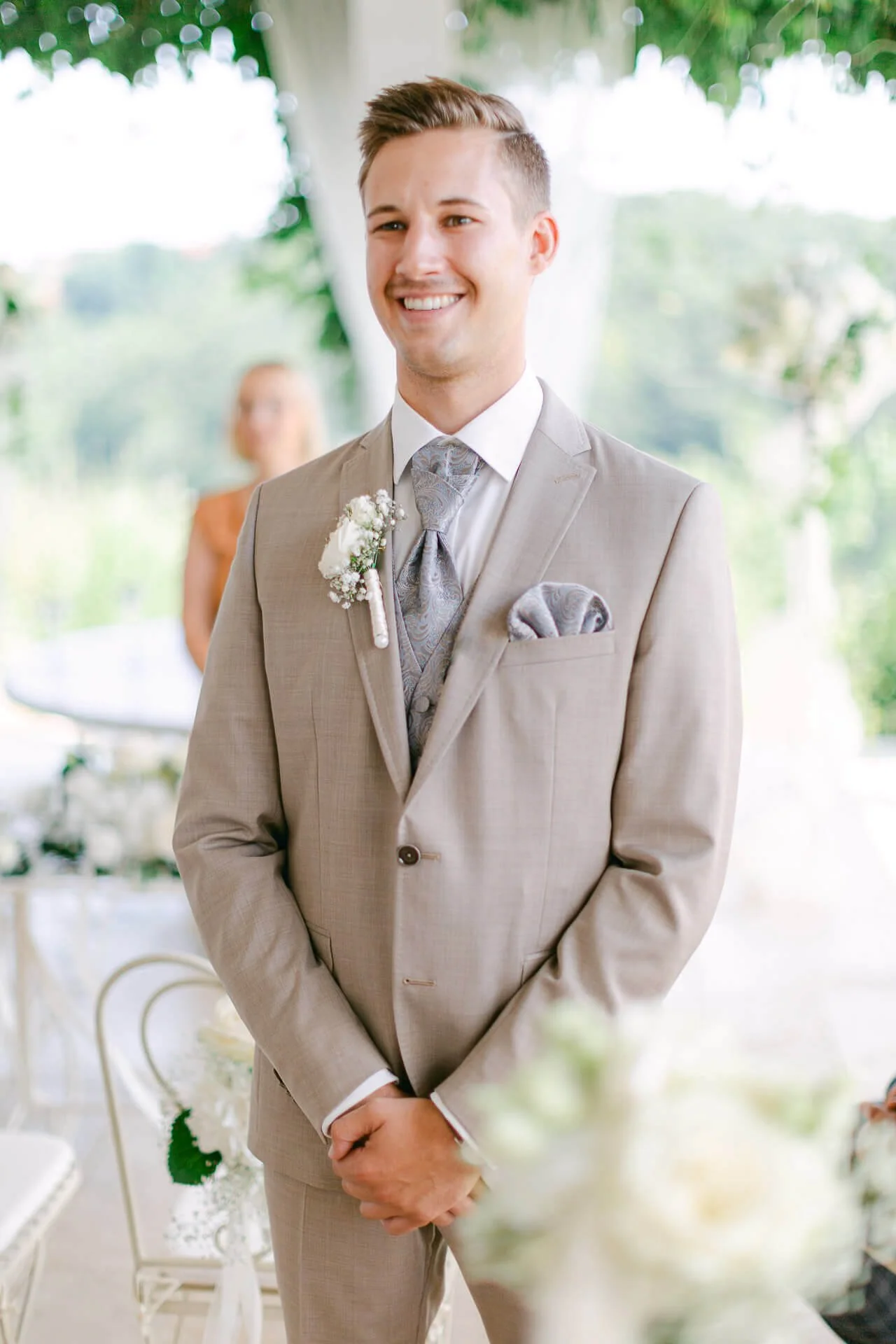 Ein lächelnder Mann in einem eleganten, beigen Anzug mit einer weißen Blume am Revers, steht in einem hellen, dekorierten Raum. Im Hintergrund ist eine Frau unscharf zu sehen.