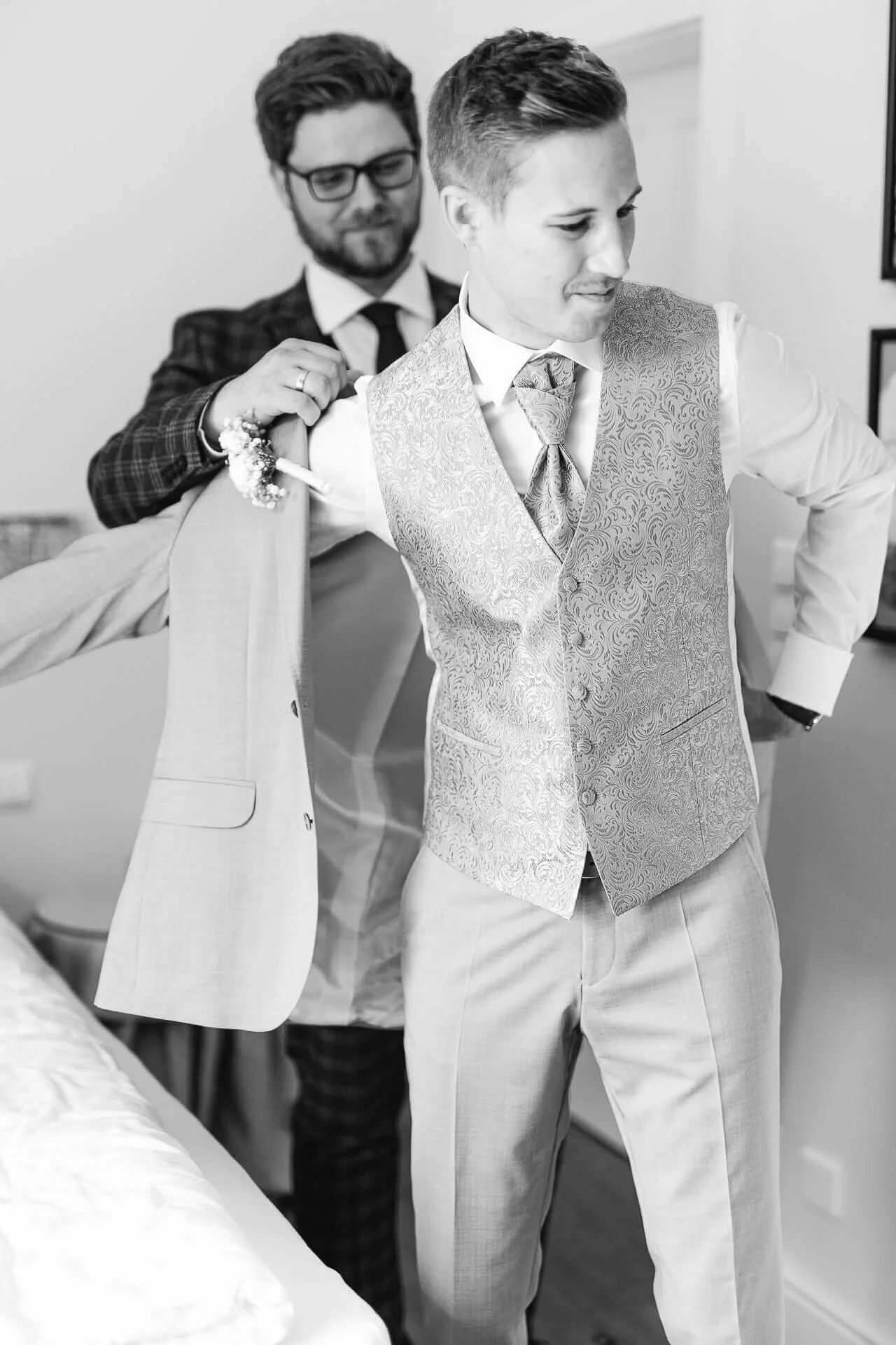 Zwei Männer in Anzügen, einer hilft dem anderen, ein Jackett anzuziehen, in einem eleganten, klassischen Stil. Schwarz-weiß Foto.