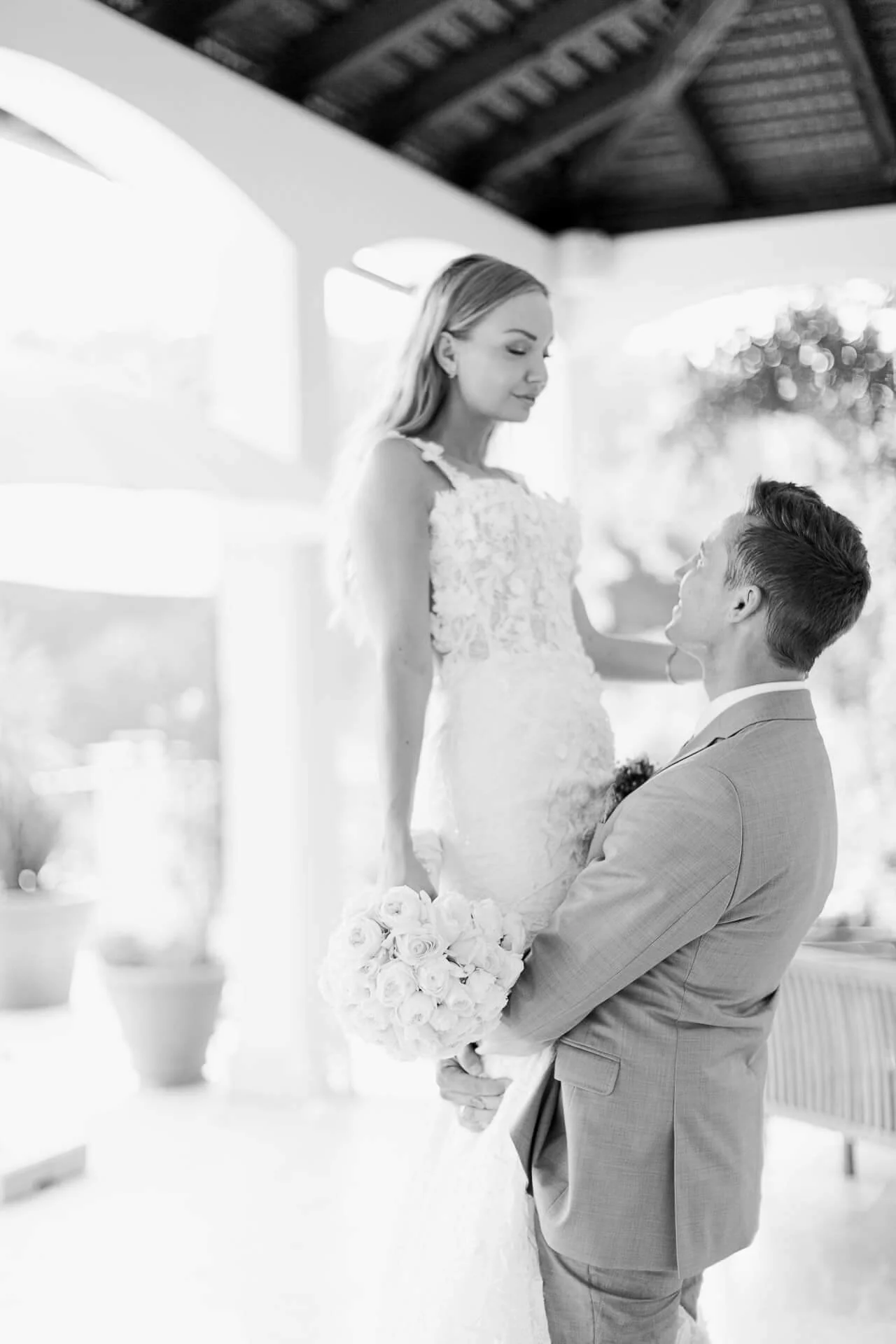 Ein Bräutigam hebt die Braut in einem Hochzeitskleid, die einen Blumenstrauß hält. Beide lächeln sich an. Schwarz-weiß Fotografie.