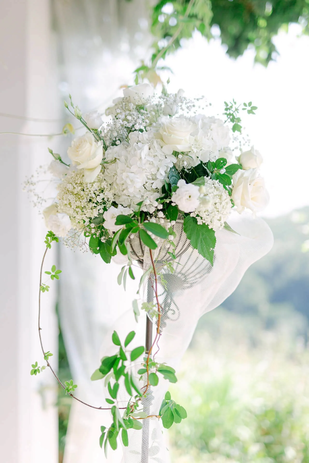 Weiße Blumenarrangement mit Rosen und Hortensien, dekoriert mit grünen Blättern, auf einem Ständer im Freien platziert, vor unscharfem Hintergrund.
