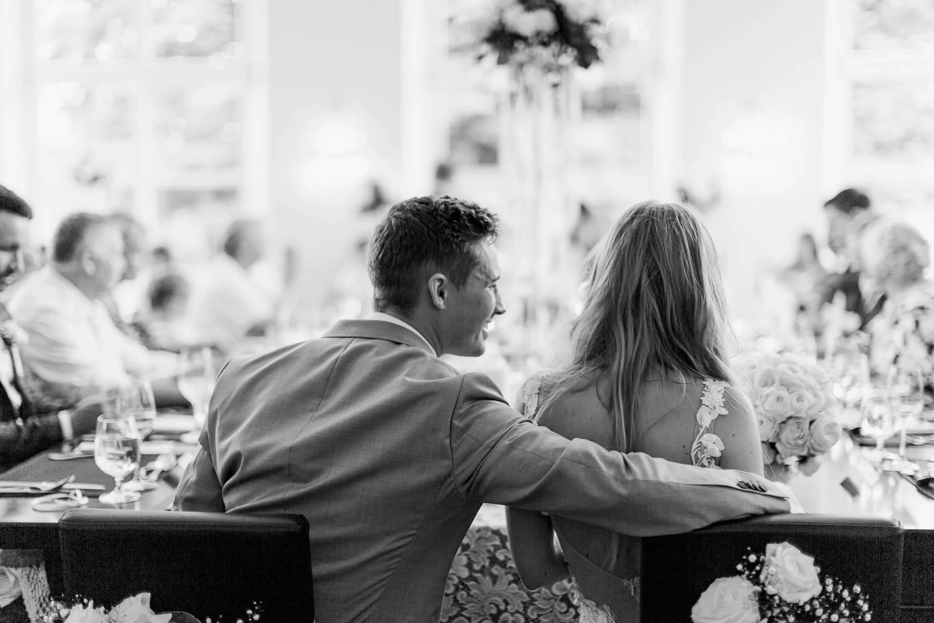 Schwarz-Weiß-Foto eines Hochzeitspaares von hinten bei einer Feier, umgeben von Gästen, mit einem Blumenarrangement auf dem Tisch.