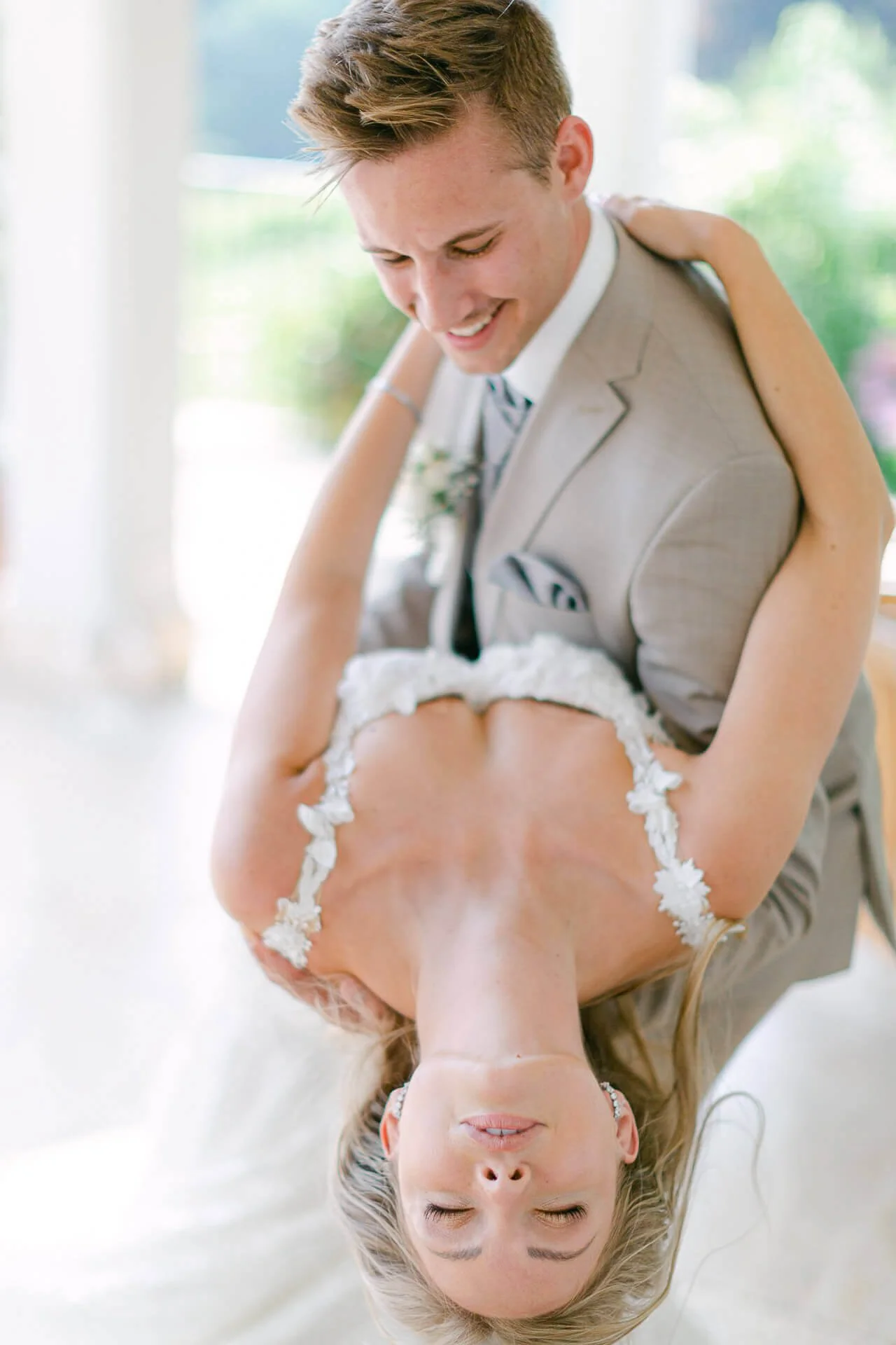 Ein Paar in Hochzeitskleidung, wobei der Mann eine Frau rückwärts hält. Die Frau trägt ein weißes Kleid und hat blondes Haar. Der Mann trägt einen beigen Anzug. Die Umgebung ist hell und natürlich.