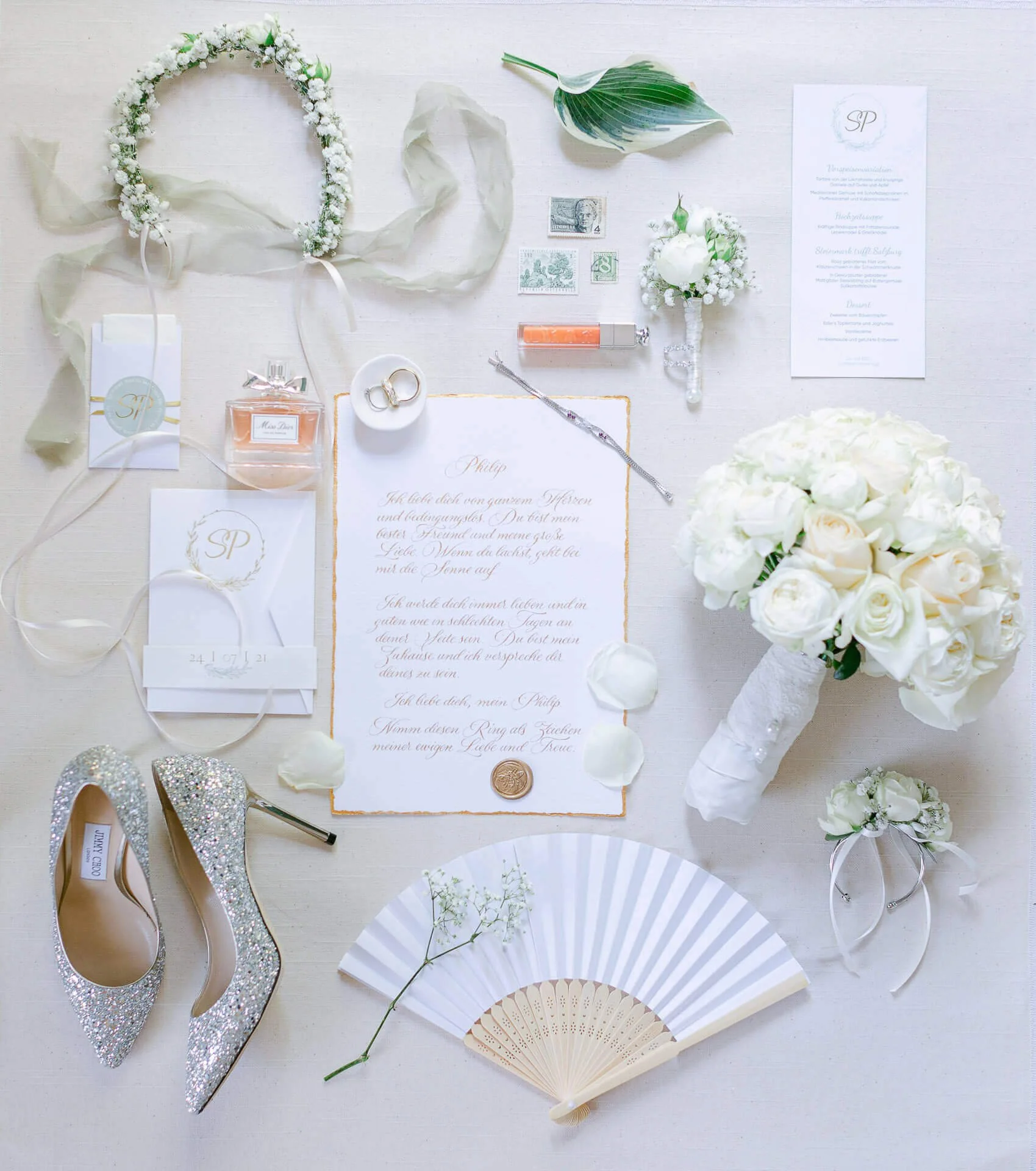 Flachlegefoto von Hochzeitsutensilien, darunter ein Blumenkranz, ein Hochzeitsstrauß aus weißen Rosen, ein paar glitzernde High Heels, ein Fächer, Parfüm, Eheringe, Hochzeitseinladung, antike Briefmarken, ein kleines Fläschchen, und ein kleines Blume