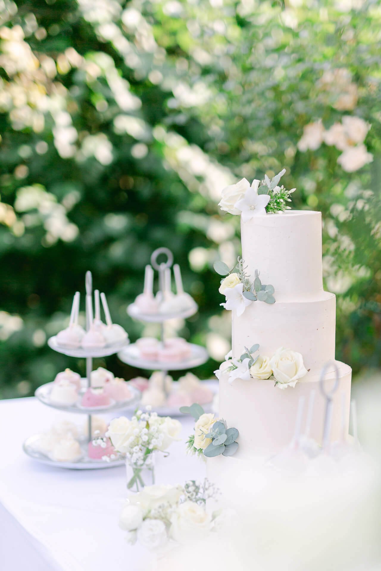 Eine dreistöckige Hochzeitstorte, verziert mit weißen Blumen, steht auf einem Tisch. Daneben stehen Etagere mit verschiedenen Süßigkeiten. Im Hintergrund ist grüne Vegetation zu sehen.