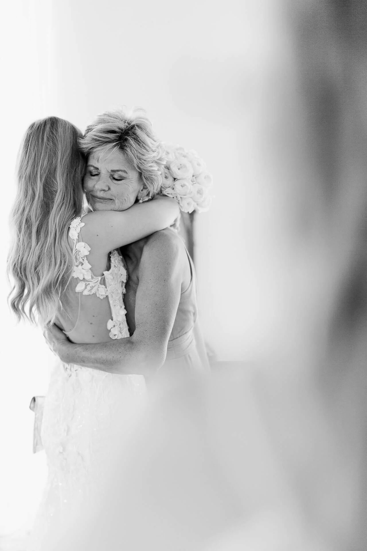 Schwarzweißfoto von zwei Frauen, die sich umarmen, eine mit langen Haaren in einem Spitzenkleid, die andere hält einen Blumenstrauß in der Hand.
