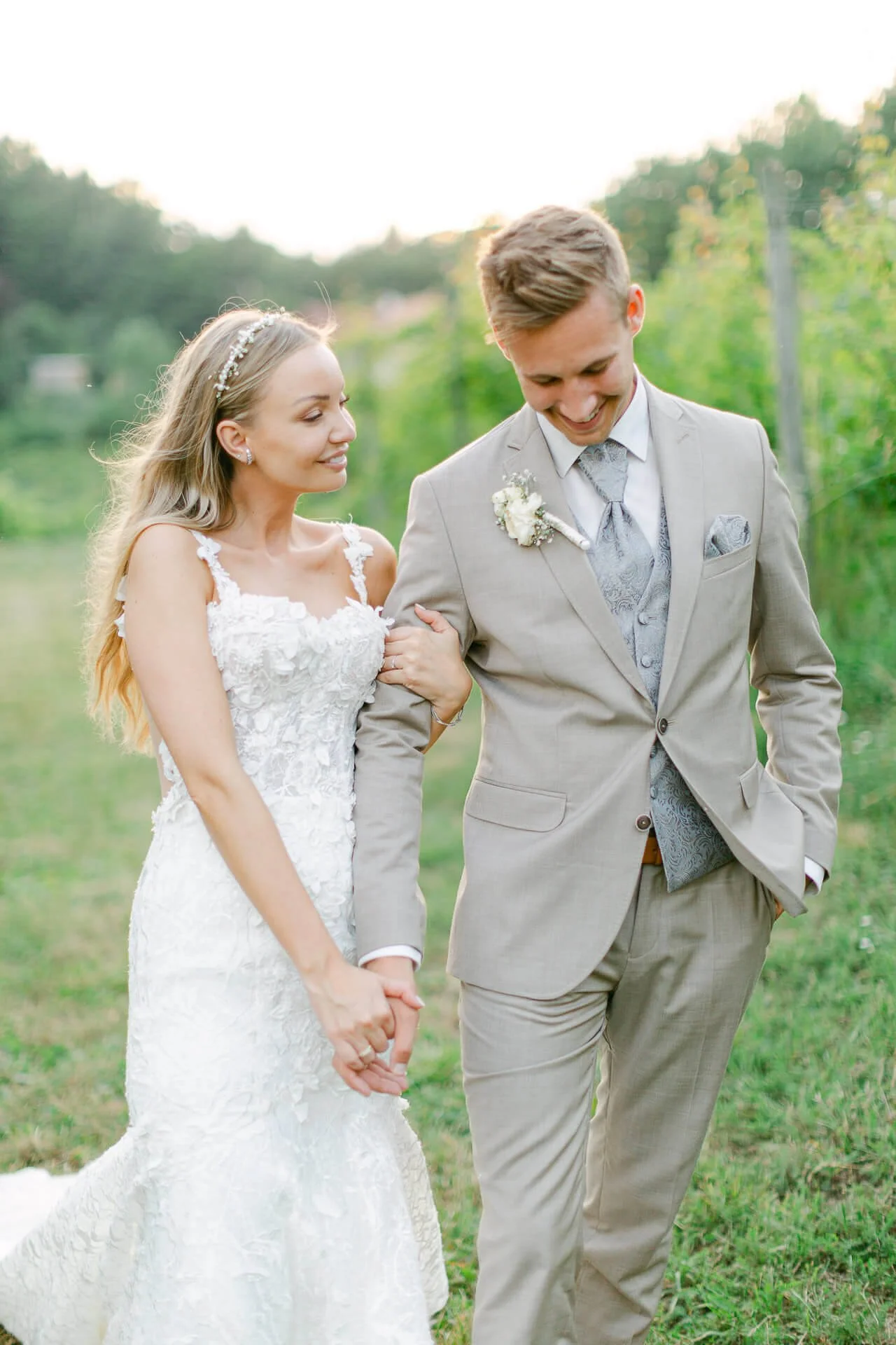 Braut und Bräutigam im Freien, die einander anlächeln und spazieren. Braut trägt ein weißes Kleid, Bräutigam einen beigefarbenen Anzug. Hintergrund von Bäumen und Wiese.