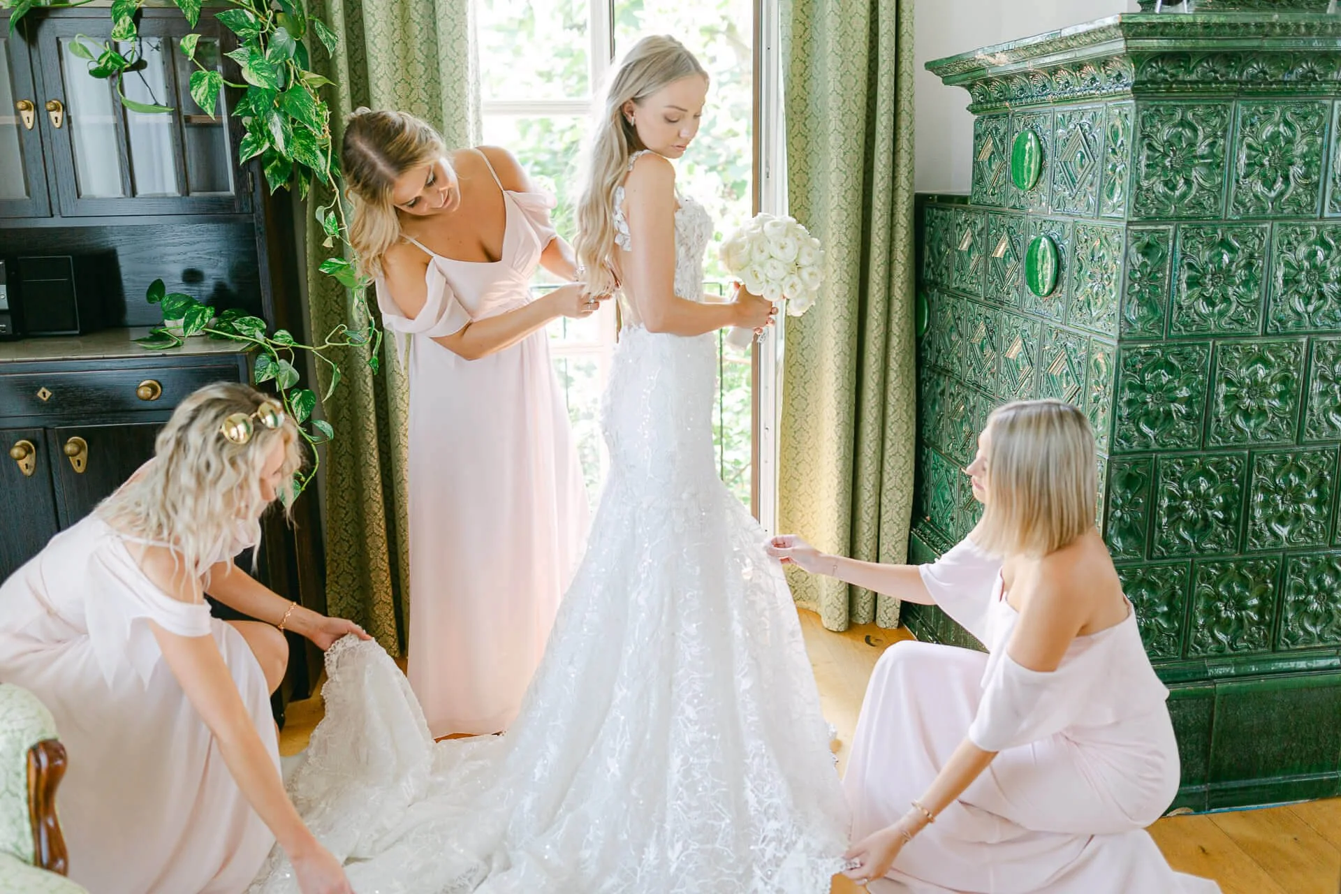 Braut in weißem Kleid mit Blumenstrauß, Assistenz von drei Frauen in Pastellkleidern, die beim Anpassen des Brautkleides helfen, vor grünem Kachelofen.